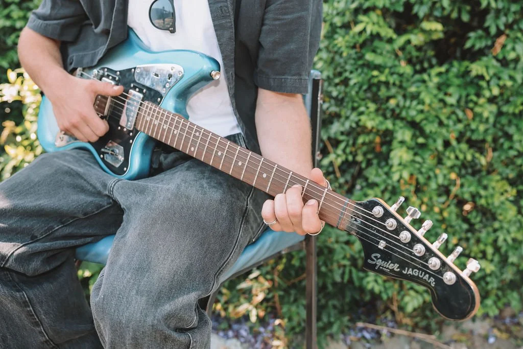 Person sitting outdoors playing a blue electric guitar with a natural green background.