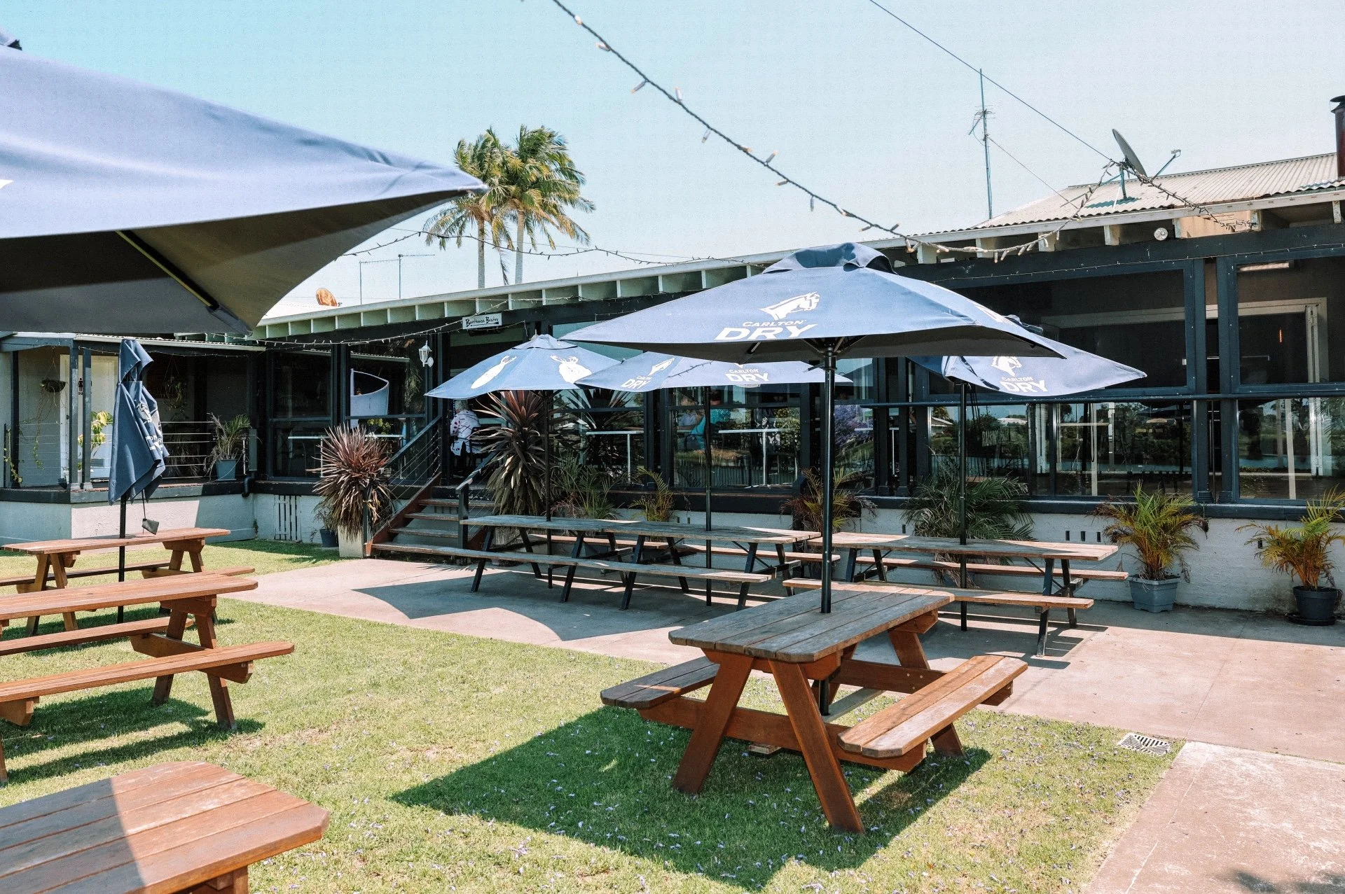 Outdoor patio area with wooden picnic tables, large umbrellas, potted plants, and a building with glass windows and a deck under a clear sky.