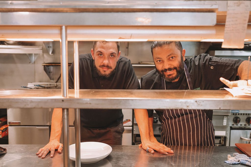 Two male chefs leaning over a kitchen counter, smiling at the camera, with kitchen appliances in the background.