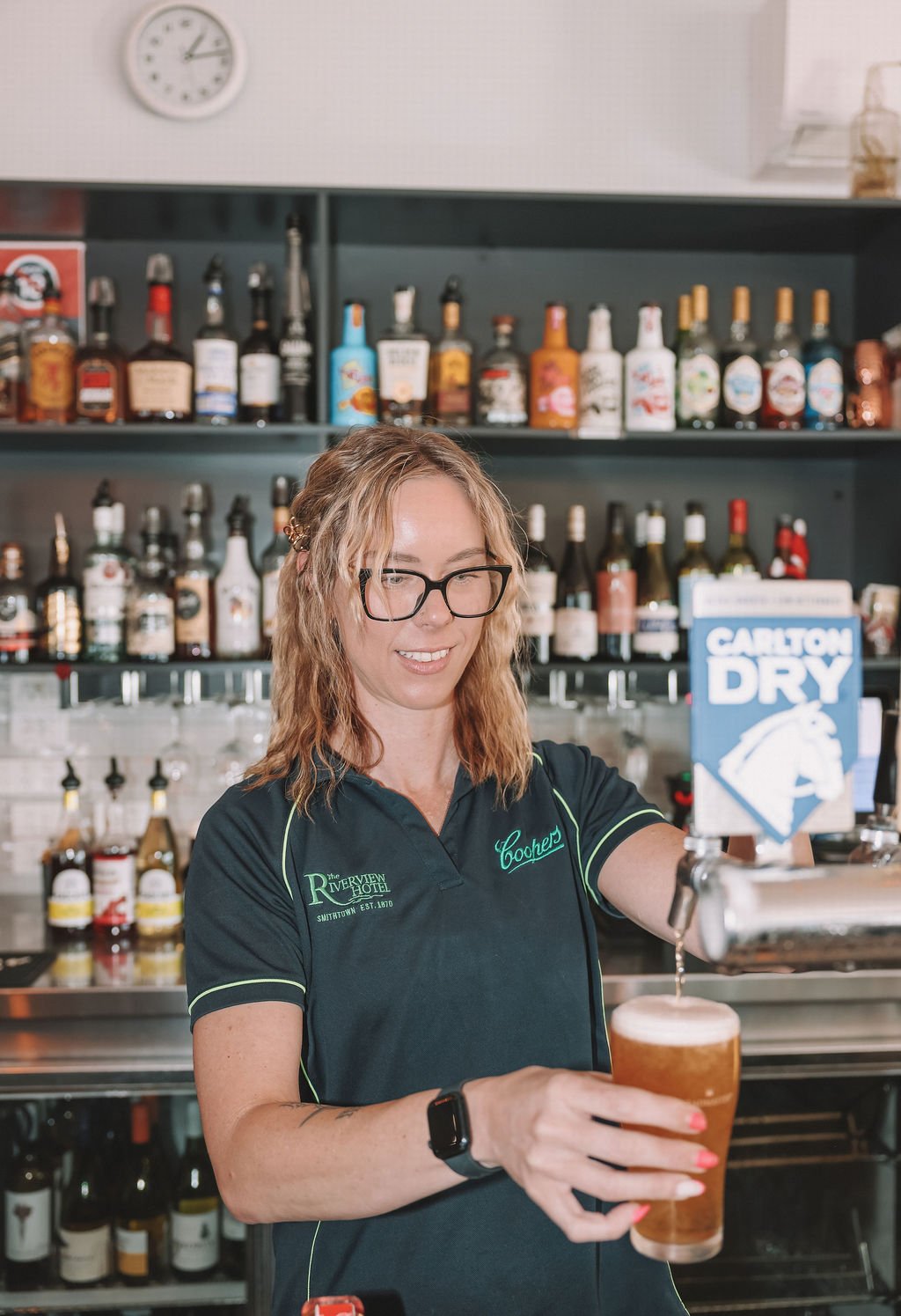 A woman with glasses and wavy blonde hair pouring a beer from a tap into a glass. She is wearing a black shirt with 'Coopers' embroidered on it and a smartwatch on her wrist. Behind her is a bar filled with various liquor bottles and a sign that says