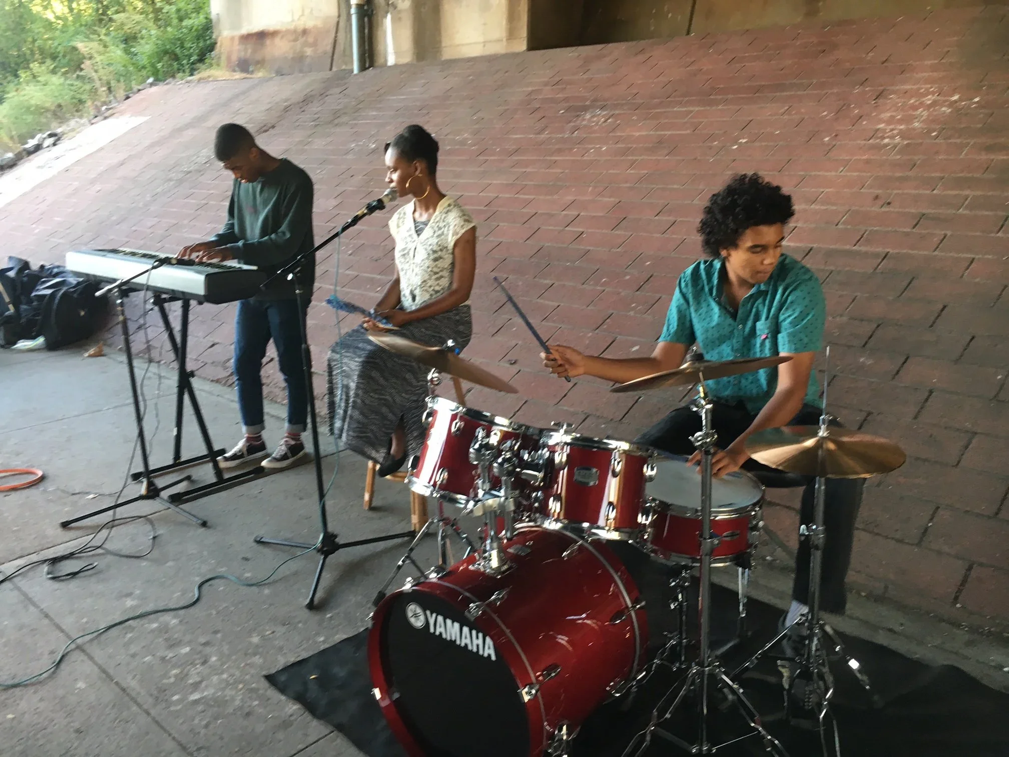 Three musicians performing outdoors on a brick-paved area. One person is playing keyboard, another singing into a microphone, and a third is sitting behind a drum set. Public Art Fellows 2017