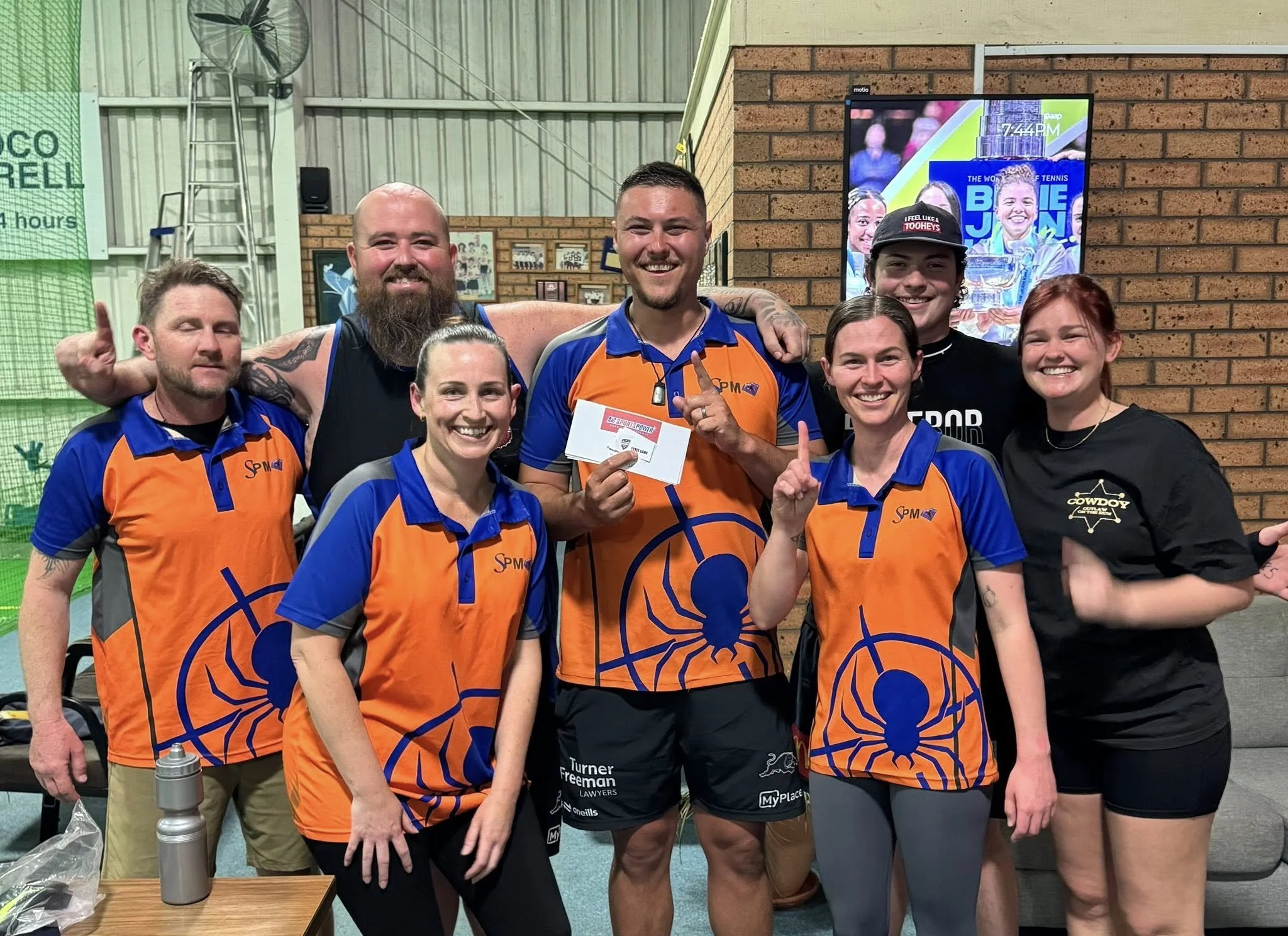 Group of seven people smiling, posing for a photo at a sports event. Five are wearing orange and blue sports jerseys with a spider logo, one is wearing a black shirt and cap, and one woman is in a black t-shirt. The man in the center is holding a che