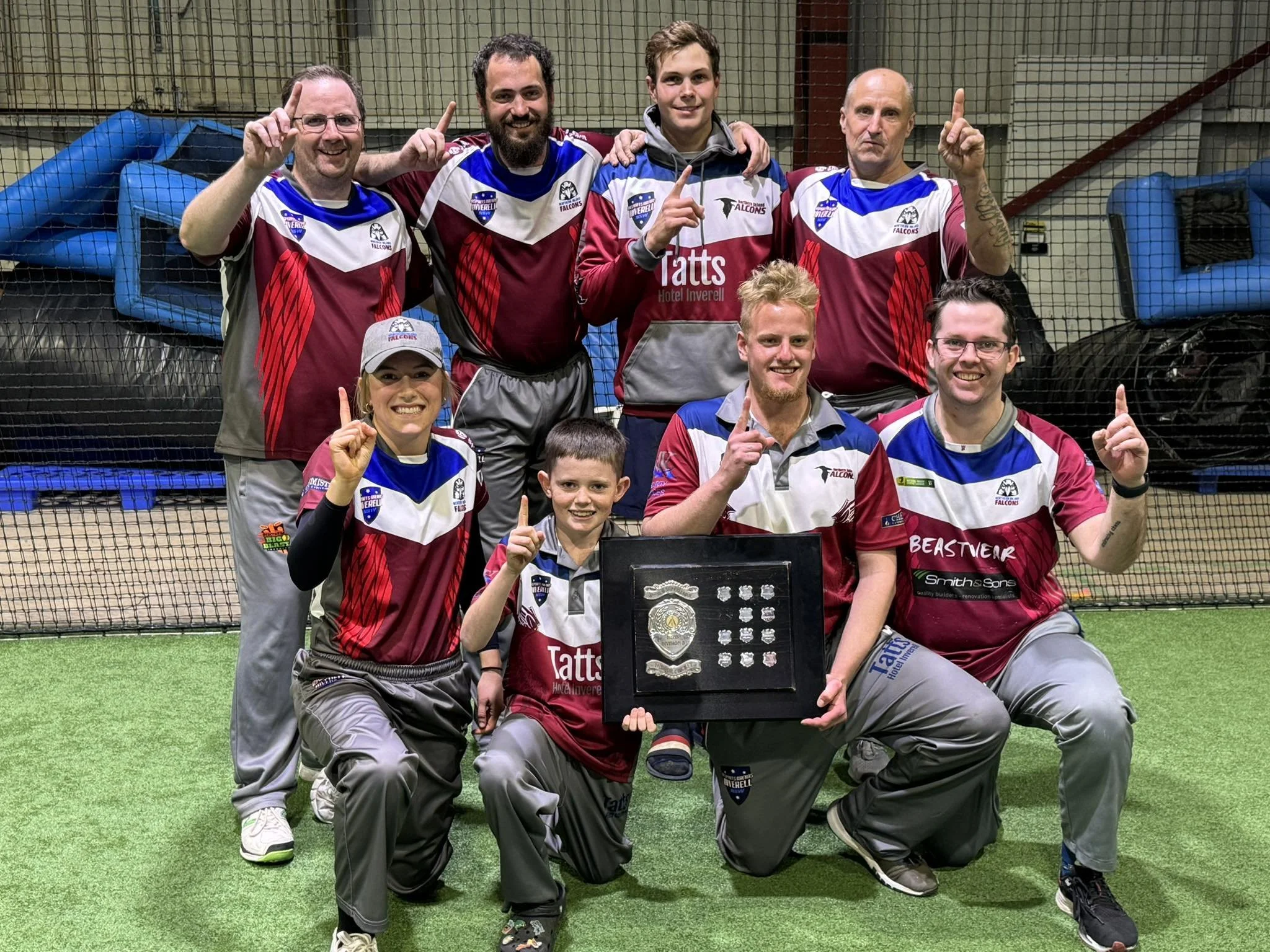A group of nine people, including seven adults and one child, posing on an indoor sports field, all wearing matching maroon, white, and blue sports jerseys, some holding up their index finger, with a framed award or trophy in front of them.