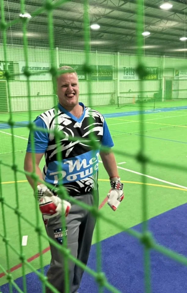 A person wearing a sports jersey and goalkeeper gloves standing inside an indoor sports facility, seen through green netting.
