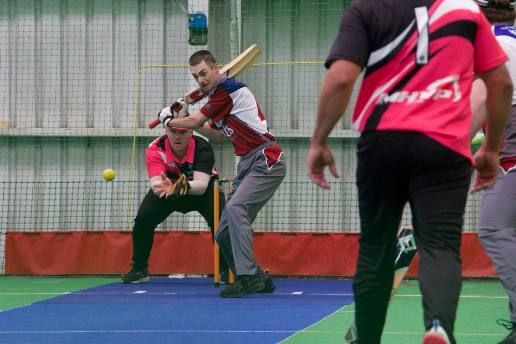 Two men in sports uniforms playing cricket indoors, one bats while the other, the wicketkeeper, is ready to catch the ball, with a yellow cricket ball in mid-air.
