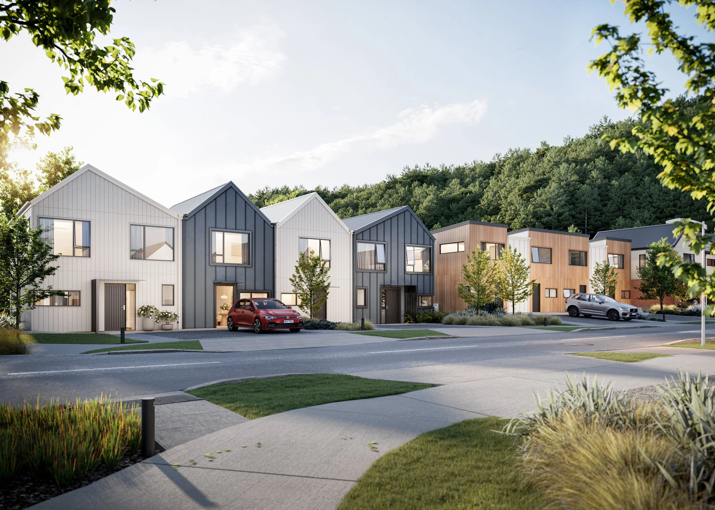 A modern residential street with two-story townhouses in various colors including white, gray, and wood finish, with cars parked in driveways, green trees, and a hillside in the background under a partly cloudy sky.