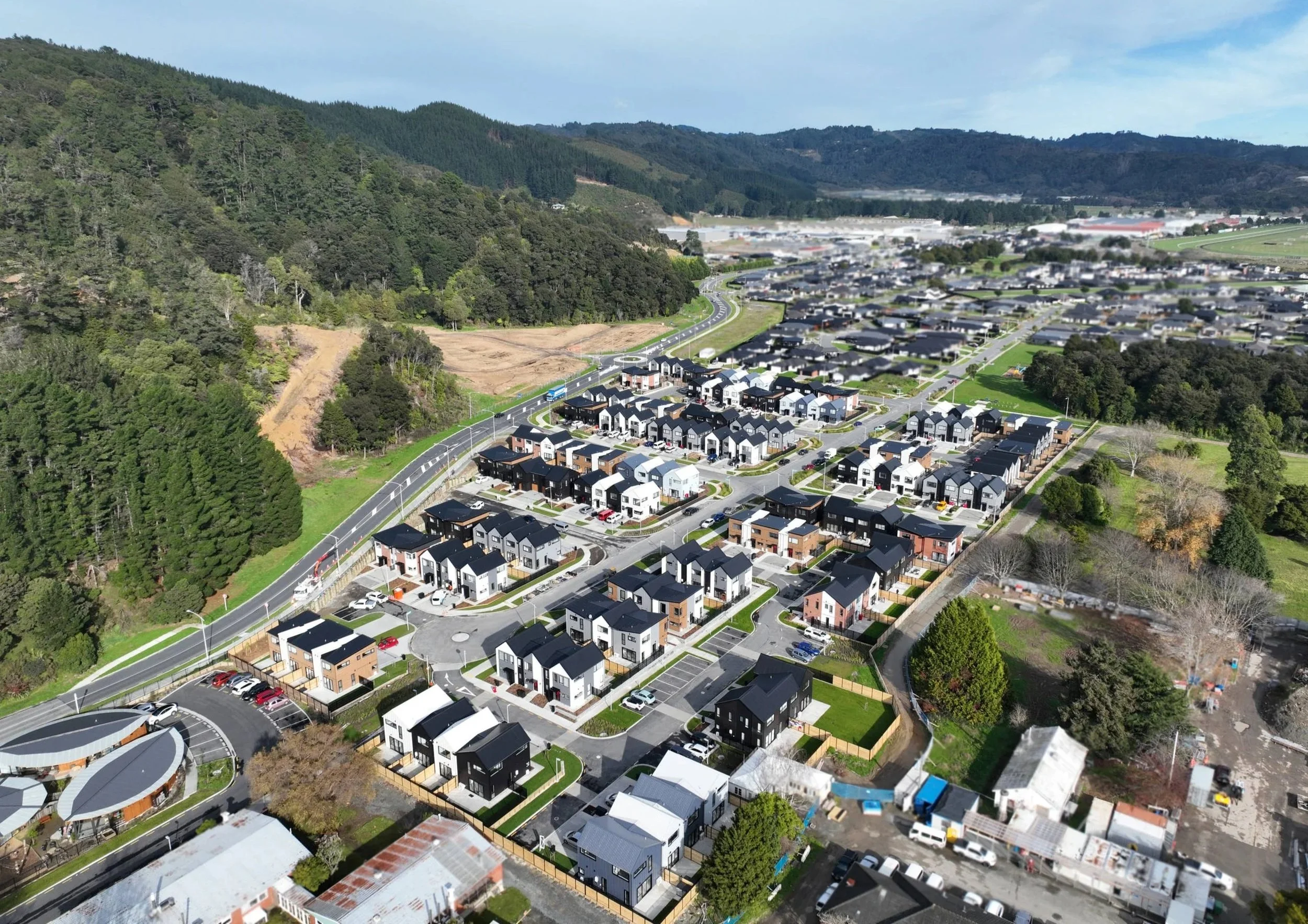 Aerial view of Urban Precinct, a Wallaceville Estate development by Gillies Group, showcases a suburban residential neighbourhood with modern houses, streets, and parking lots, surrounded by green trees and hills.