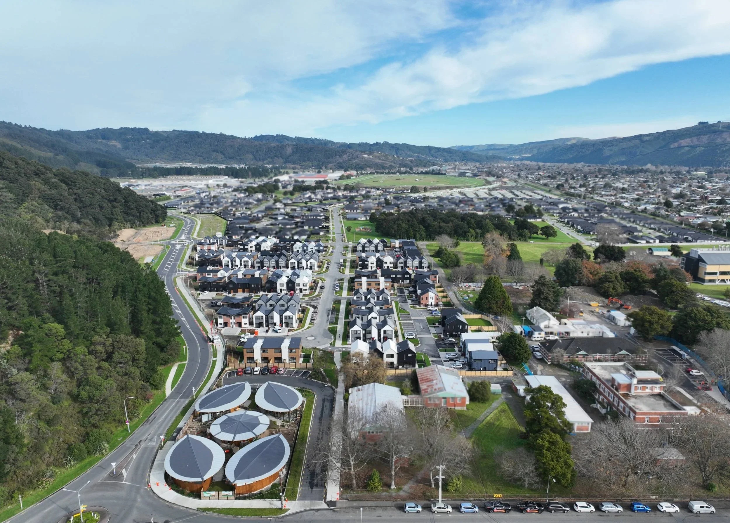 Aerial view of a suburban area with residential houses, commercial buildings, green parks, and surrounding hills under a partly cloudy sky.