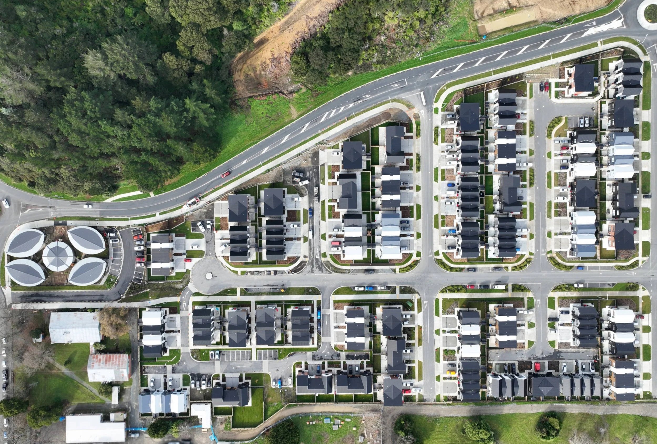 An aerial view of a residential neighbourhood with houses, roads, and parked cars. A forested area is visible at the top left.