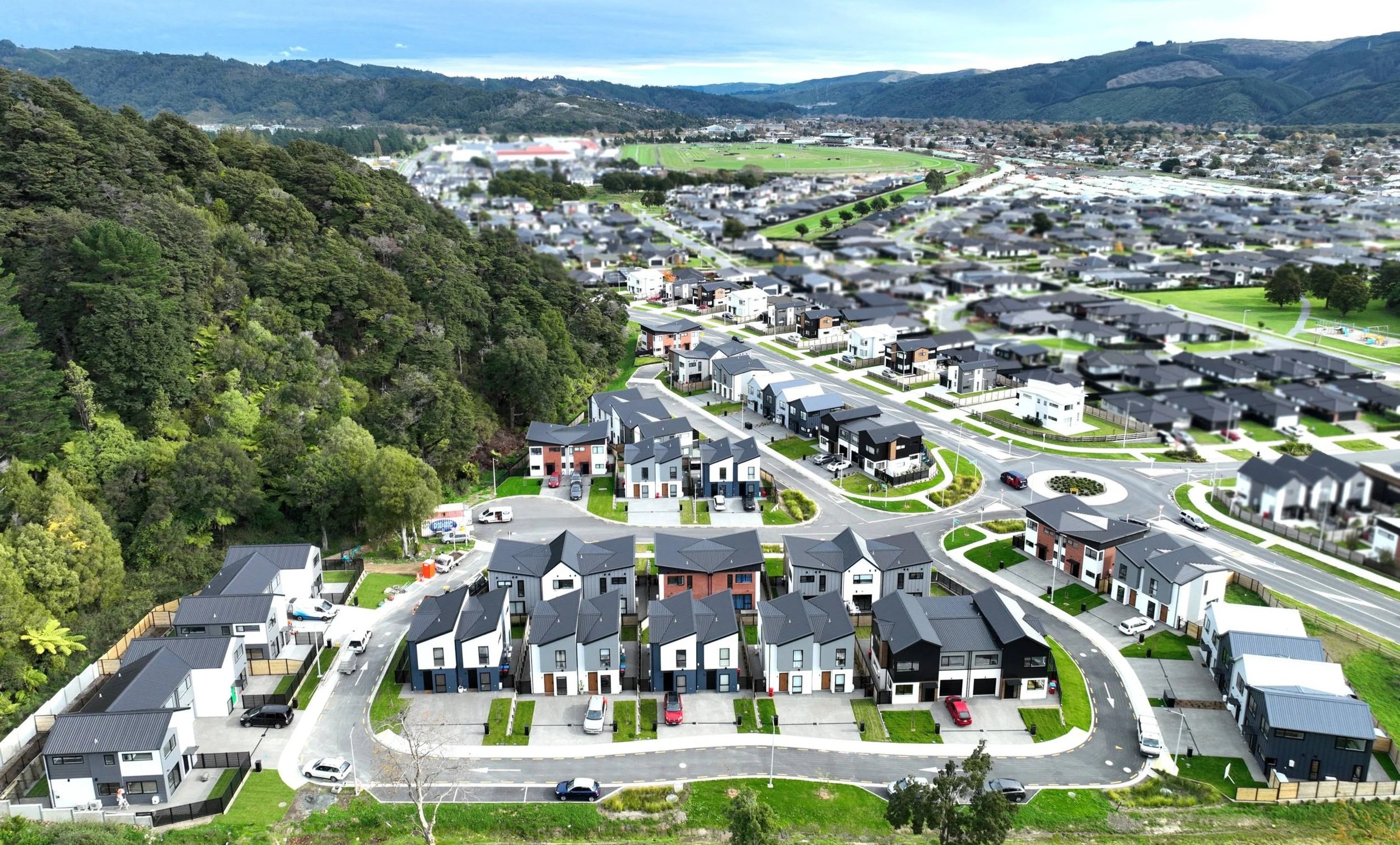 Aerial view of a suburban neighbourhood with modern houses, parking lots, and winding roads near a wooded hillside, with distant mountains and a sports field in the background.