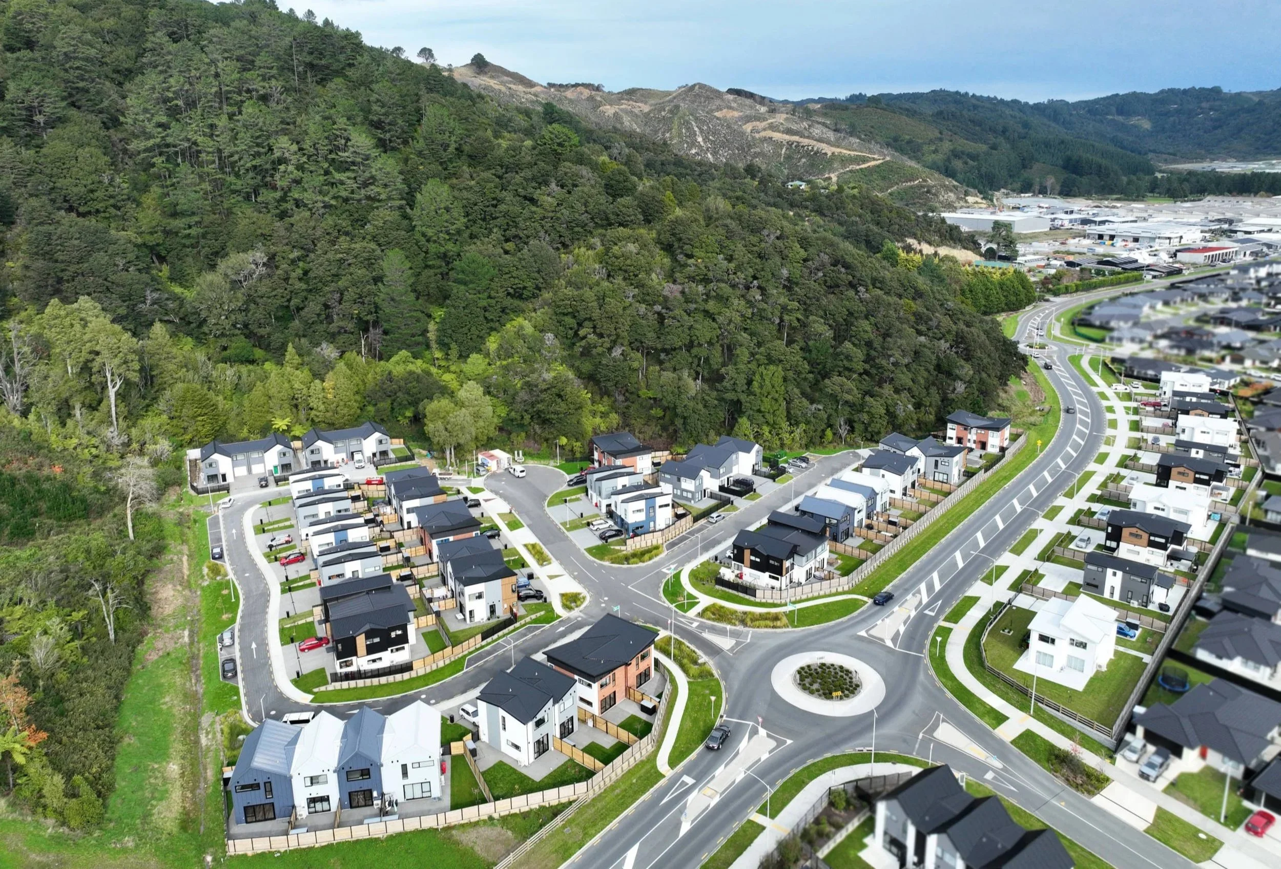 Aerial view of a residential neighbourhood with modern white and black houses, green lawns, and a circular roundabout, located adjacent to a forested hill and industrial area in the distance.