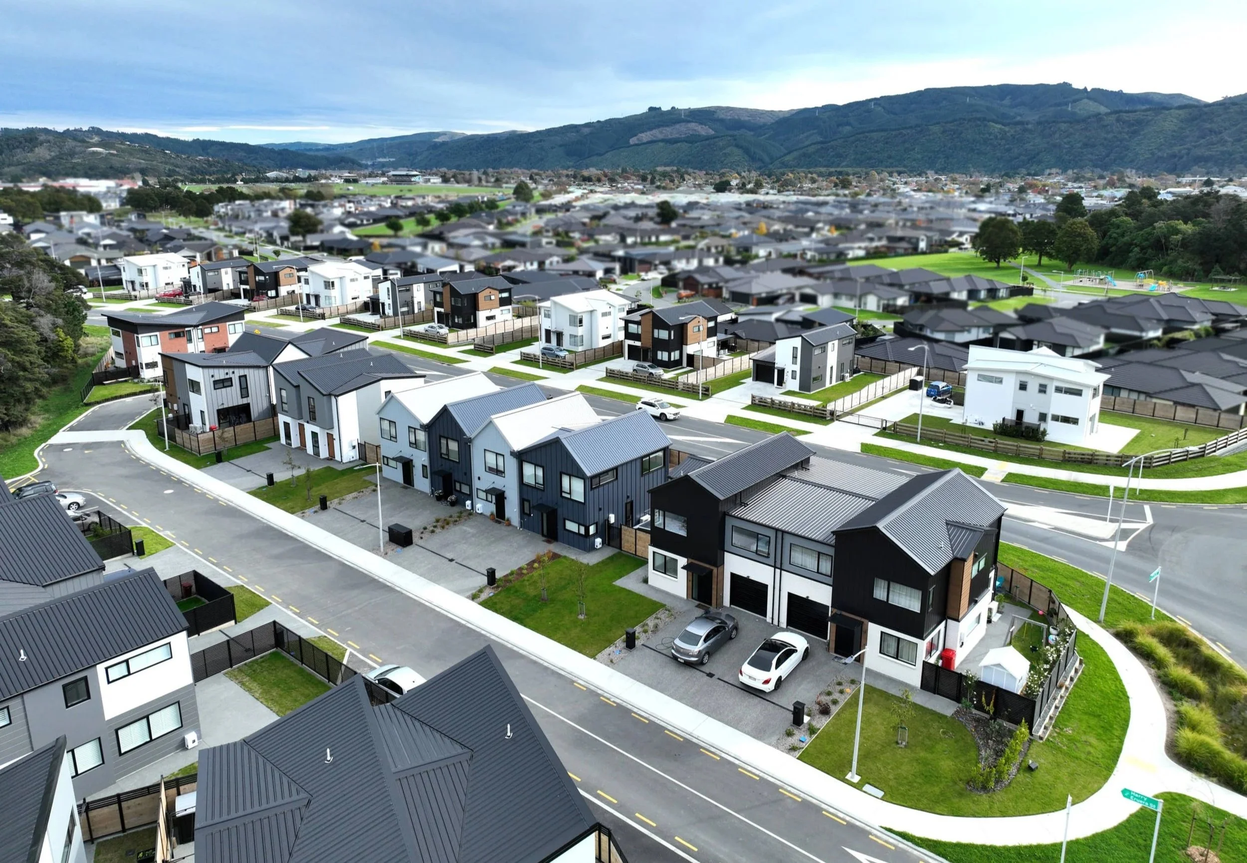 Aerial view of a modern residential neighbourhood with contemporary houses, roads, green lawns, and mountains in the background.