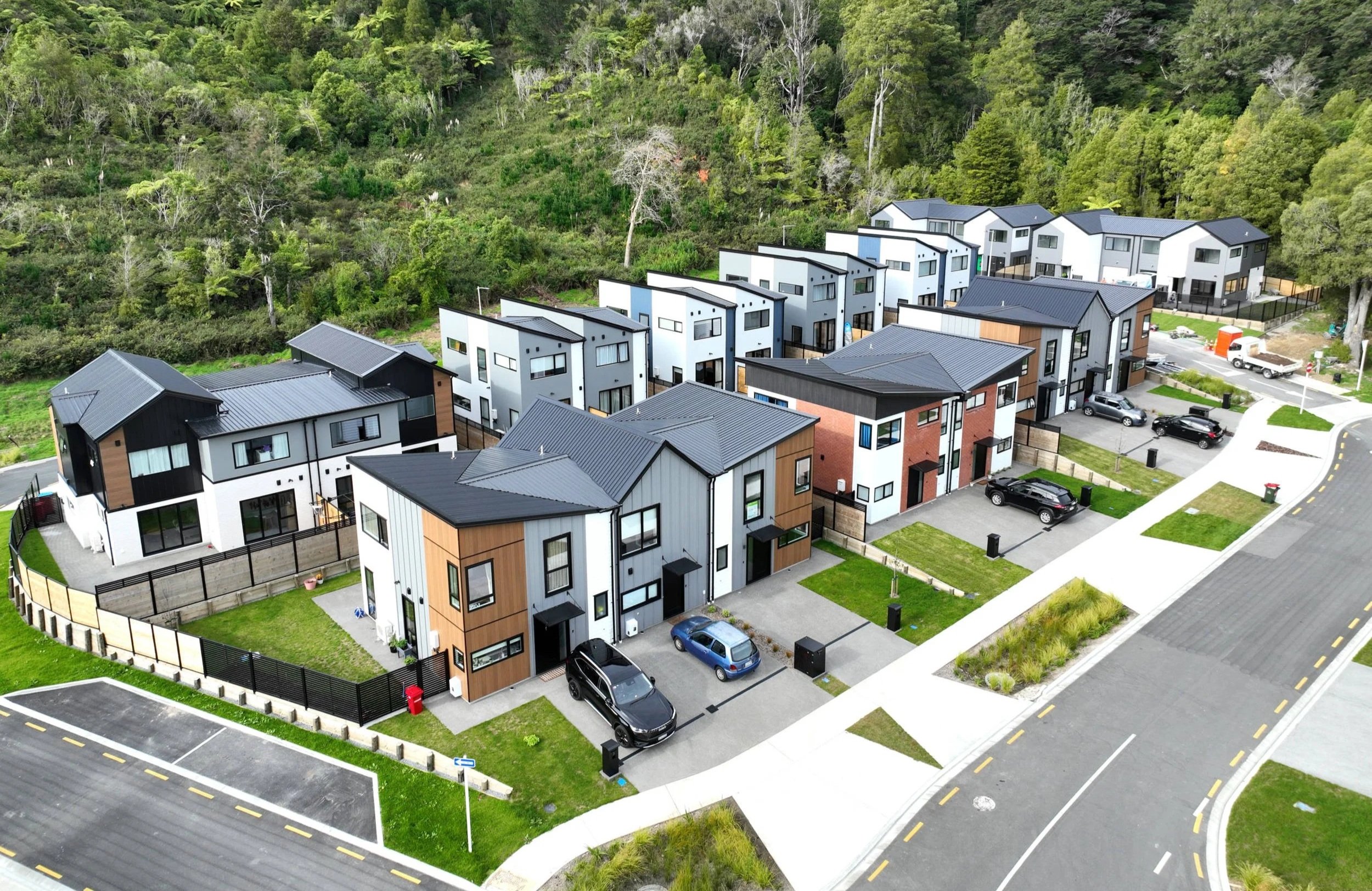 Modern residential housing development with standalone homes and townhouses, parking spaces, and landscaped green areas, situated near a forested hillside.