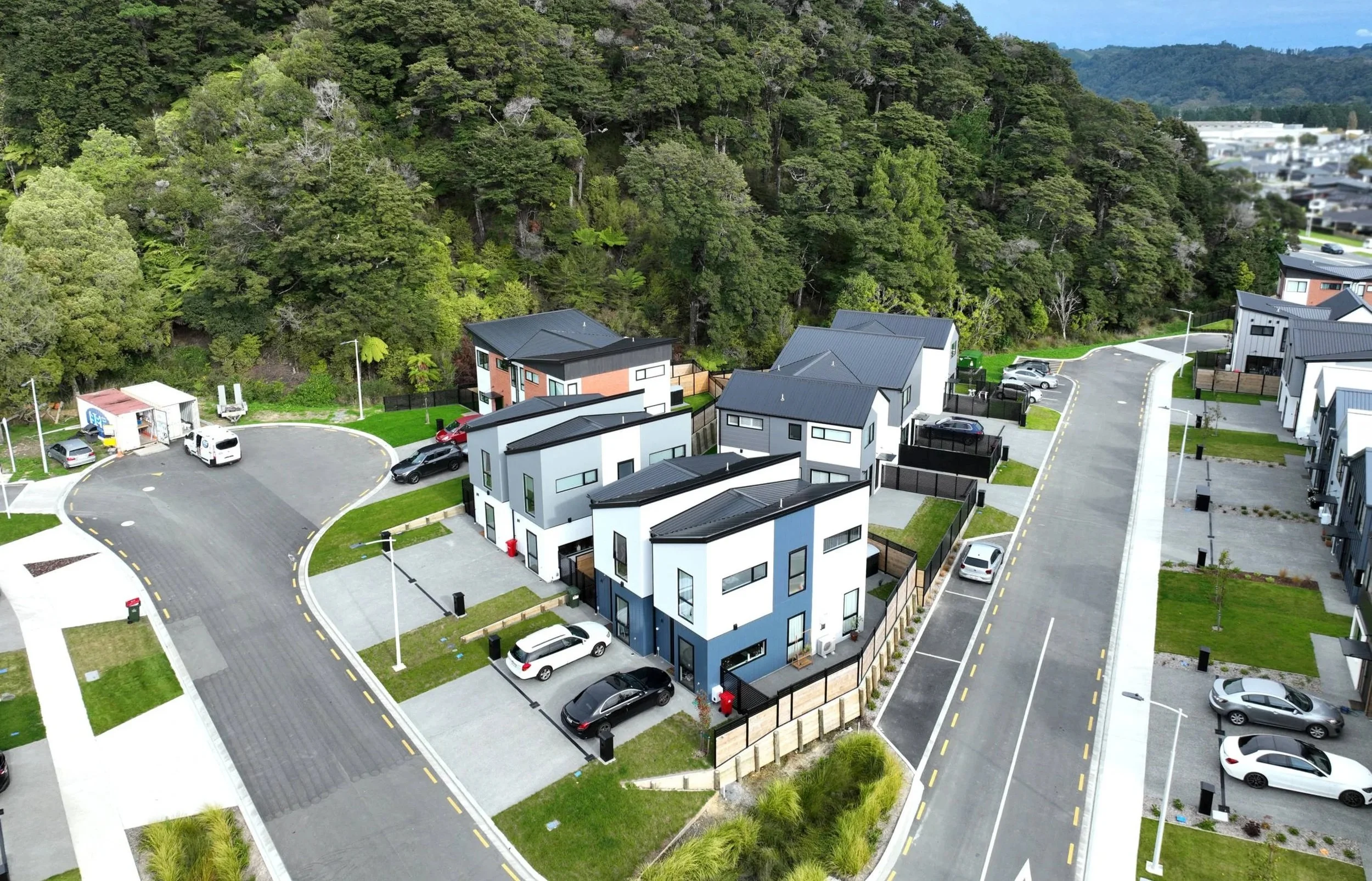 Aerial view of a modern residential neighbourhood with white and gray houses, driveways, parked cars, and a wooded hillside in the background.
