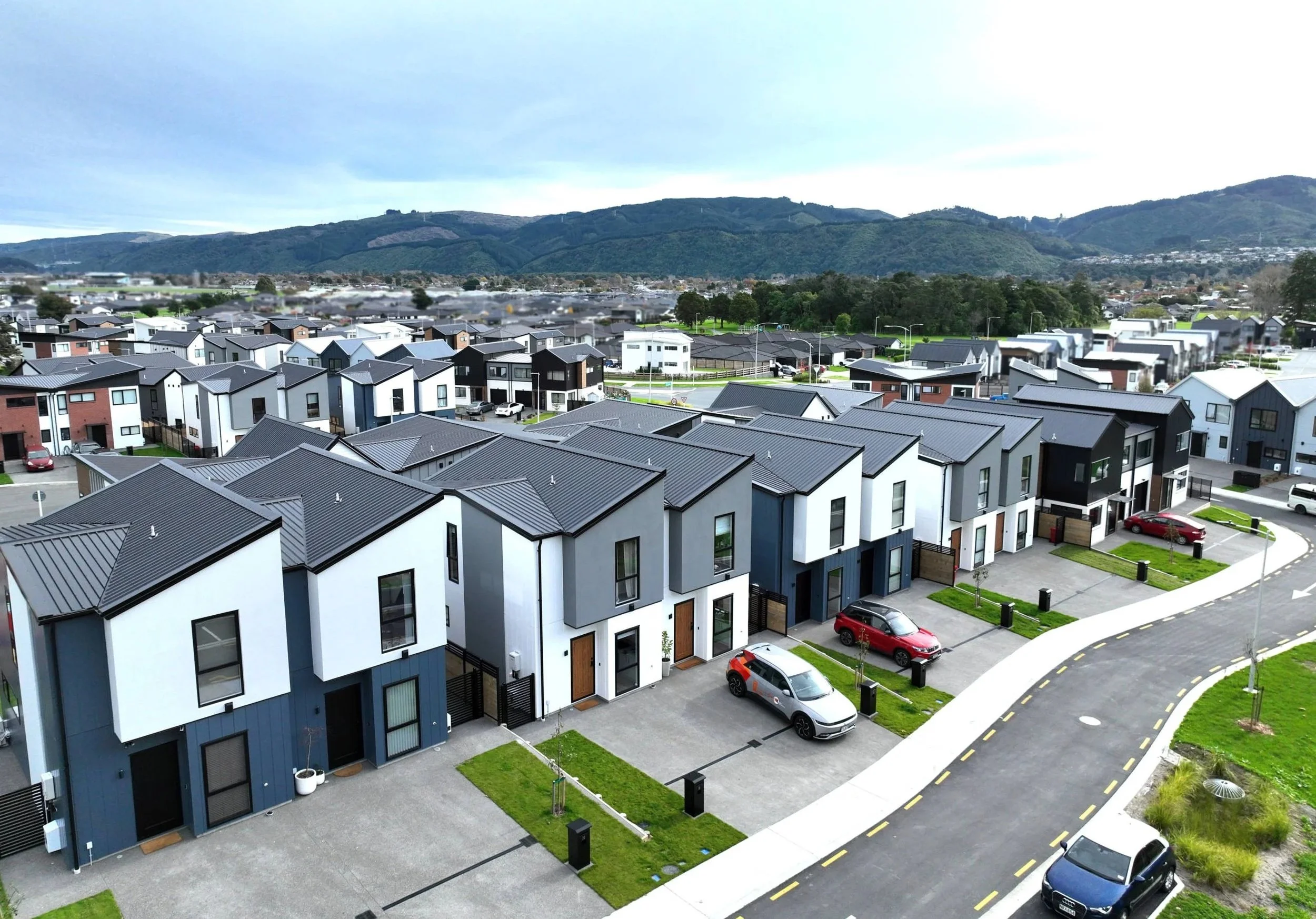 Drone photo of Urban Precinct in Wallaceville Estate, a development by Gillies Group, shows a view of modern residential neighbourhood with townhouses, parked cars, and landscaping against distant hills and cloudy sky.