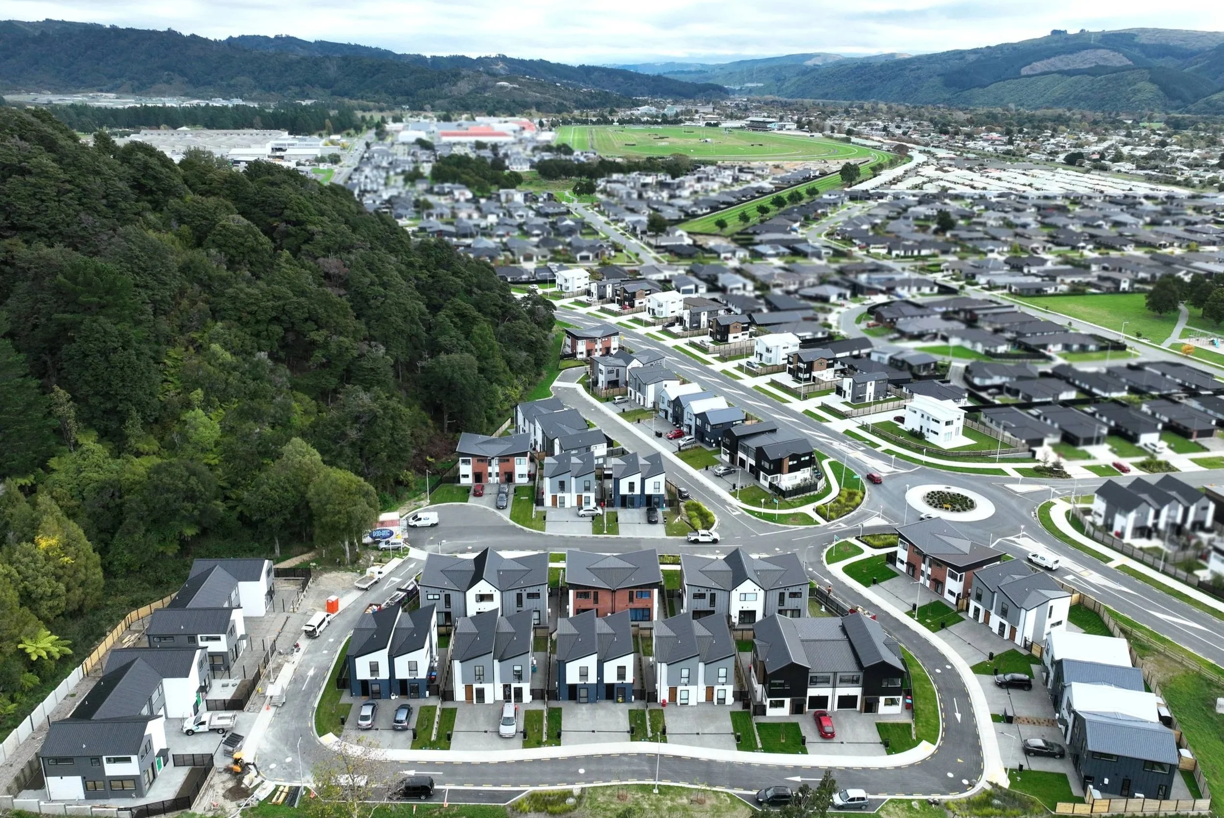 Aerial view of a modern residential neighbourhood with houses, streets, and cars, adjacent to a green, wooded hillside.