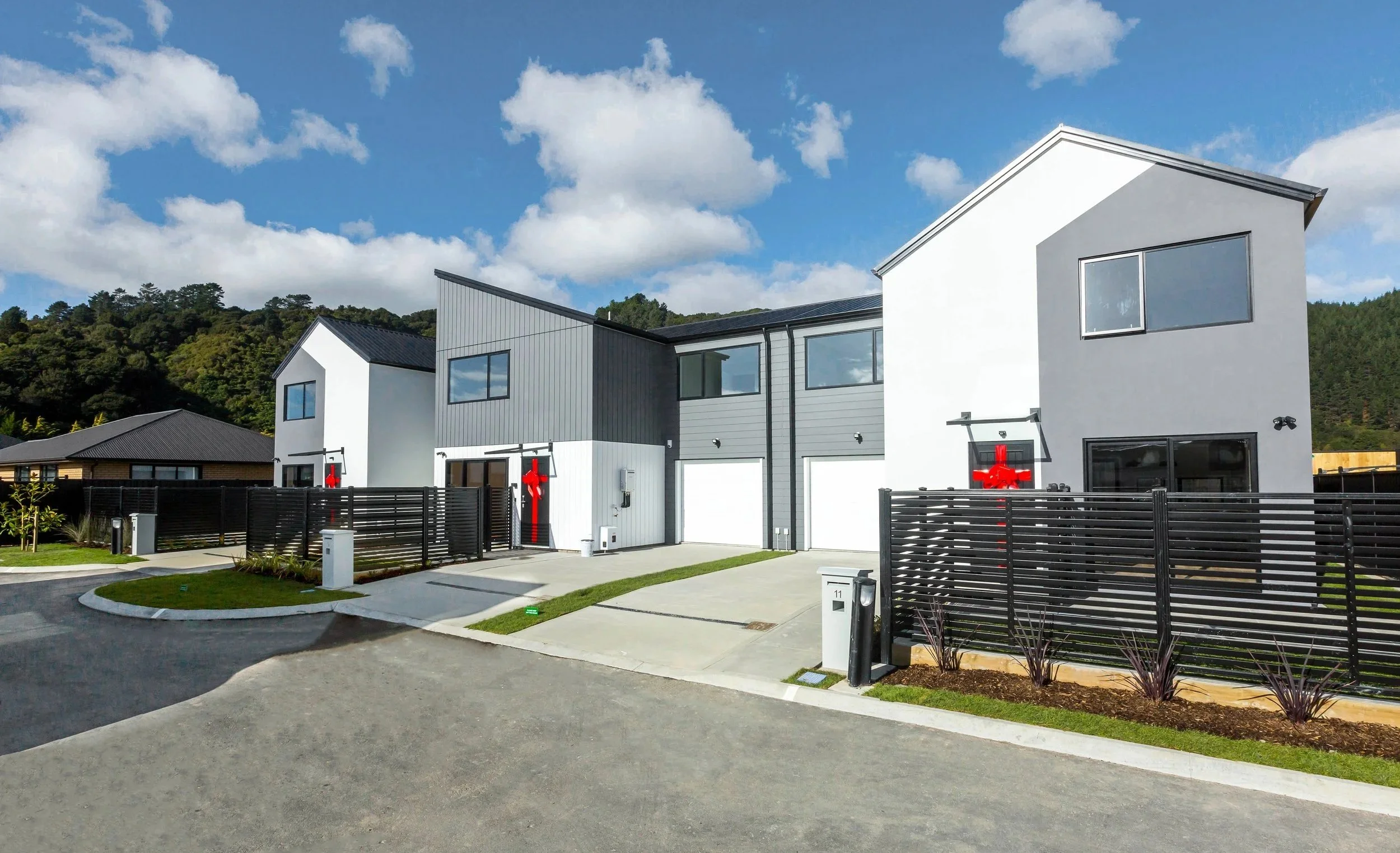 Type E houses from The Reserve: Final Release, with gray and white exteriors, black fences, and decorative red bows on the doors, set against a backdrop of green hills and a partly cloudy sky.