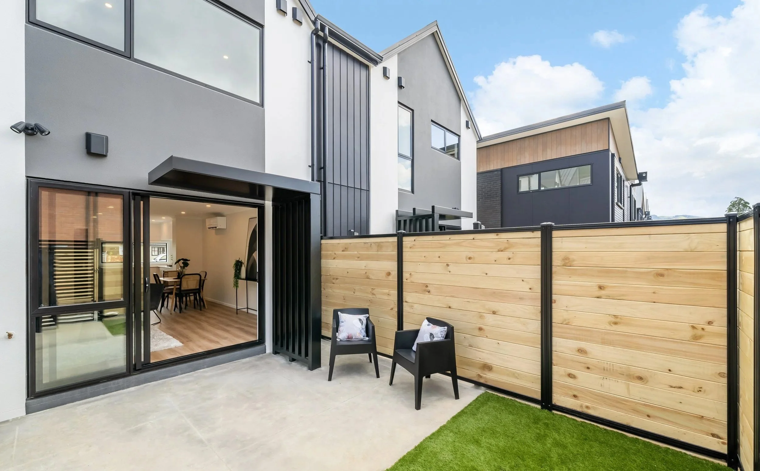 Modern townhouse backyard with concrete patio, two black chairs with pillows, wooden privacy fence, sliding glass door leading inside, and neighboring contemporary houses.