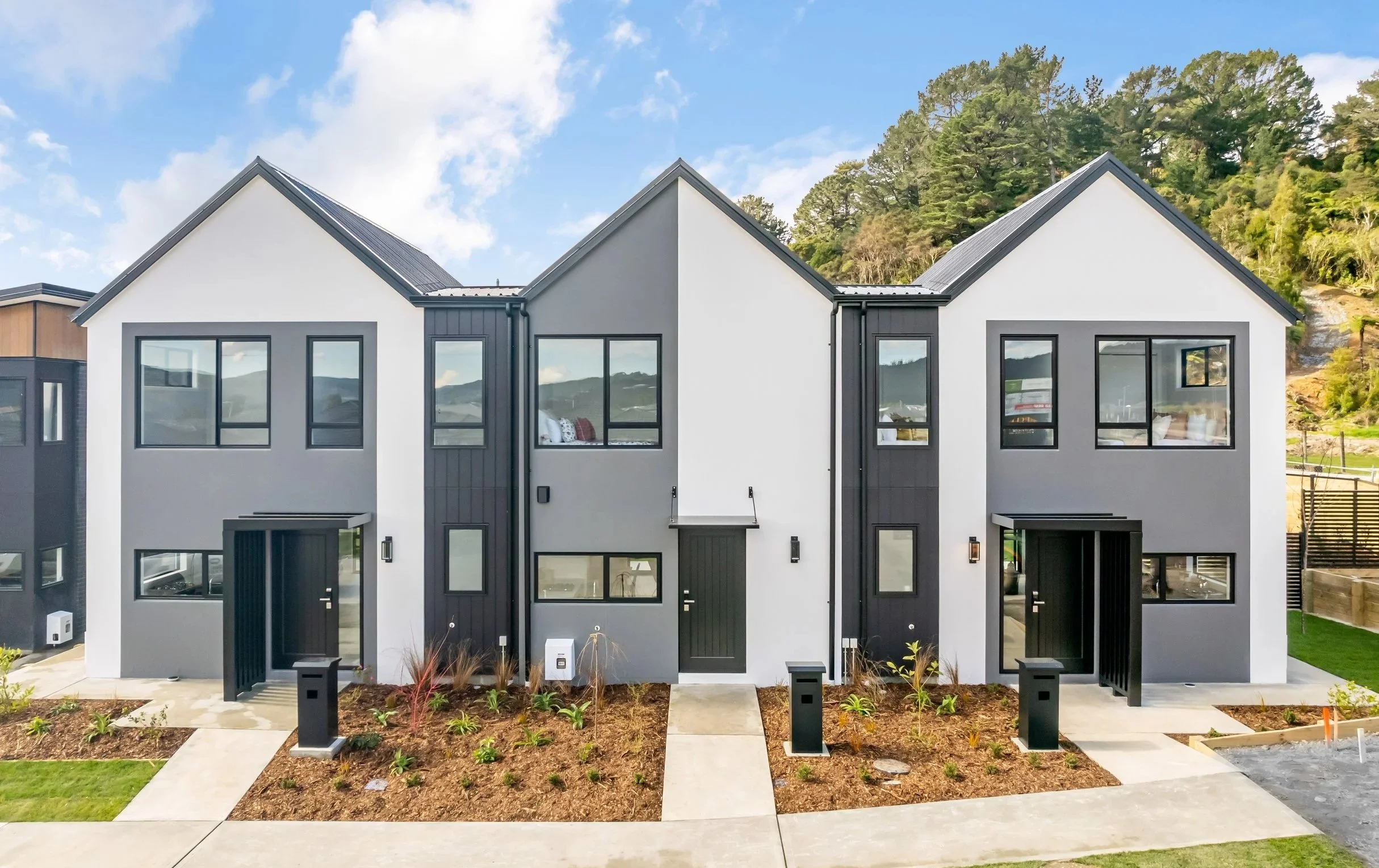 The Reserve: Final Release's Type C and Type B houses with gray and white exteriors, black trim, and landscaping in front, set against a backdrop of hills and trees on a sunny day, located in Wallaceville Estate.
