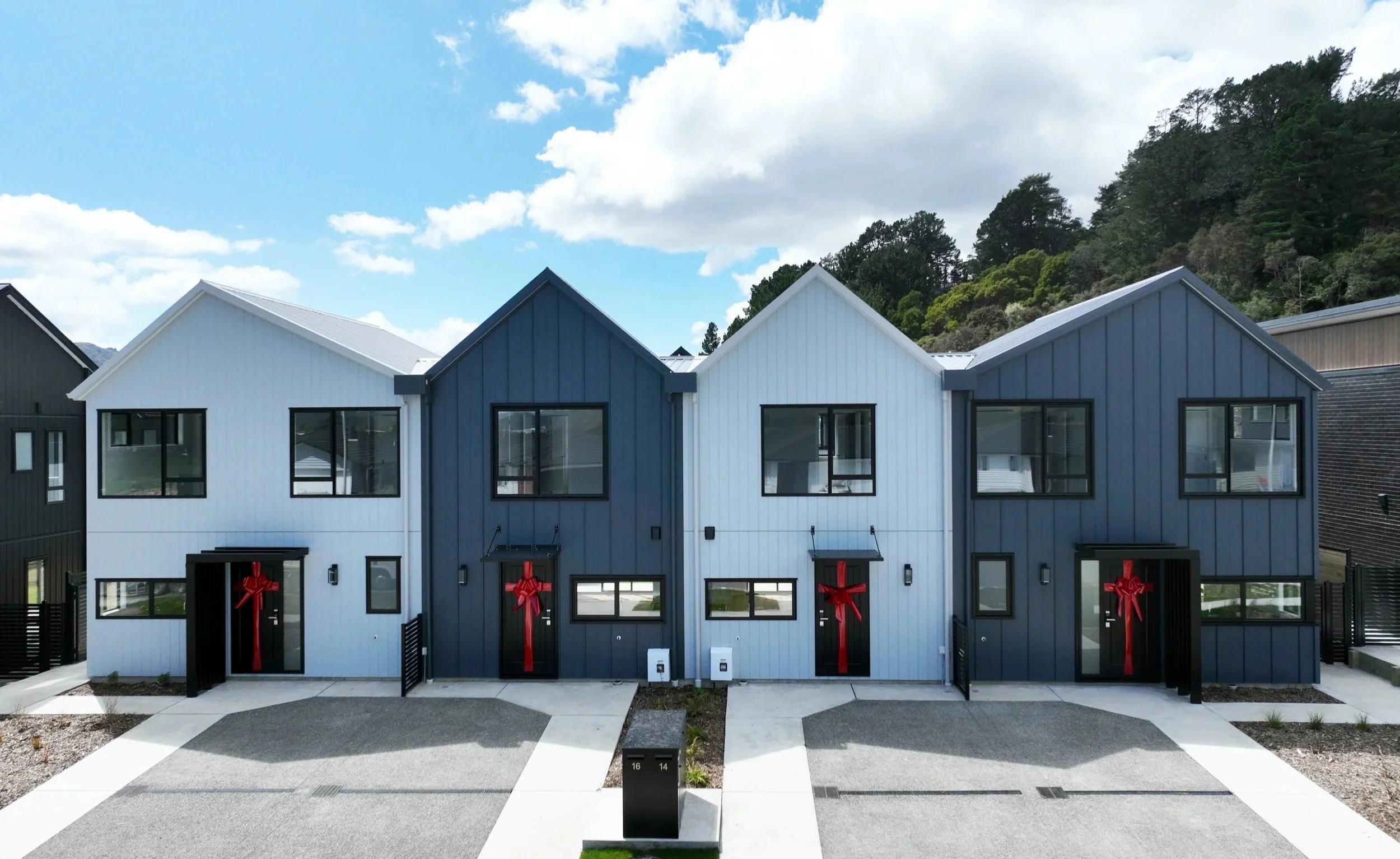 Row of Type C and Type B houses decorated with large red bows, with a paved driveway and landscaping in front, under a partly cloudy sky, located in Wallaceville Estate in Wellington.