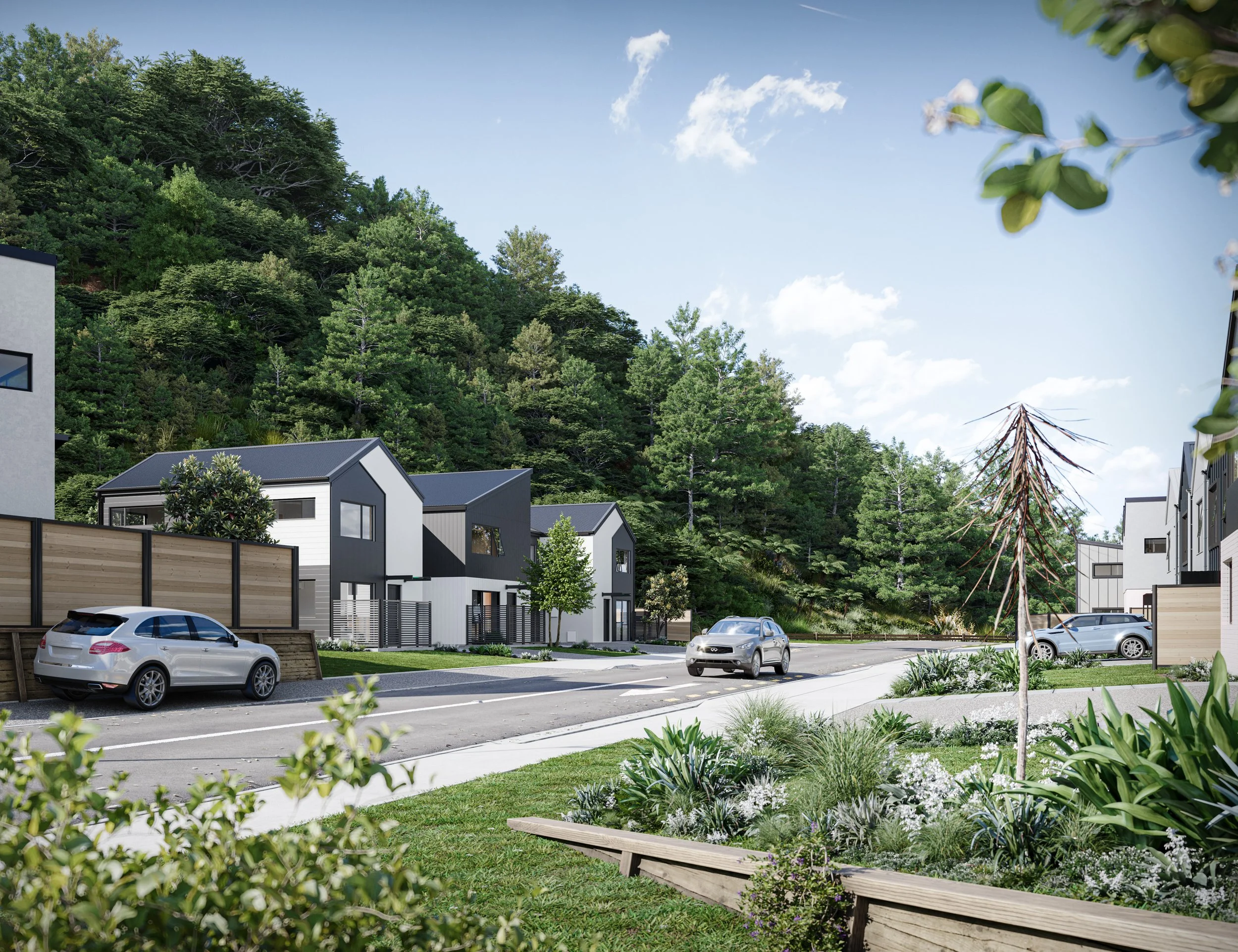A modern residential neighbourhood with multi-story houses, parked cars on a residential street, lush greenery, trees, and a wooded hillside in the background under a bright blue sky with a few clouds.