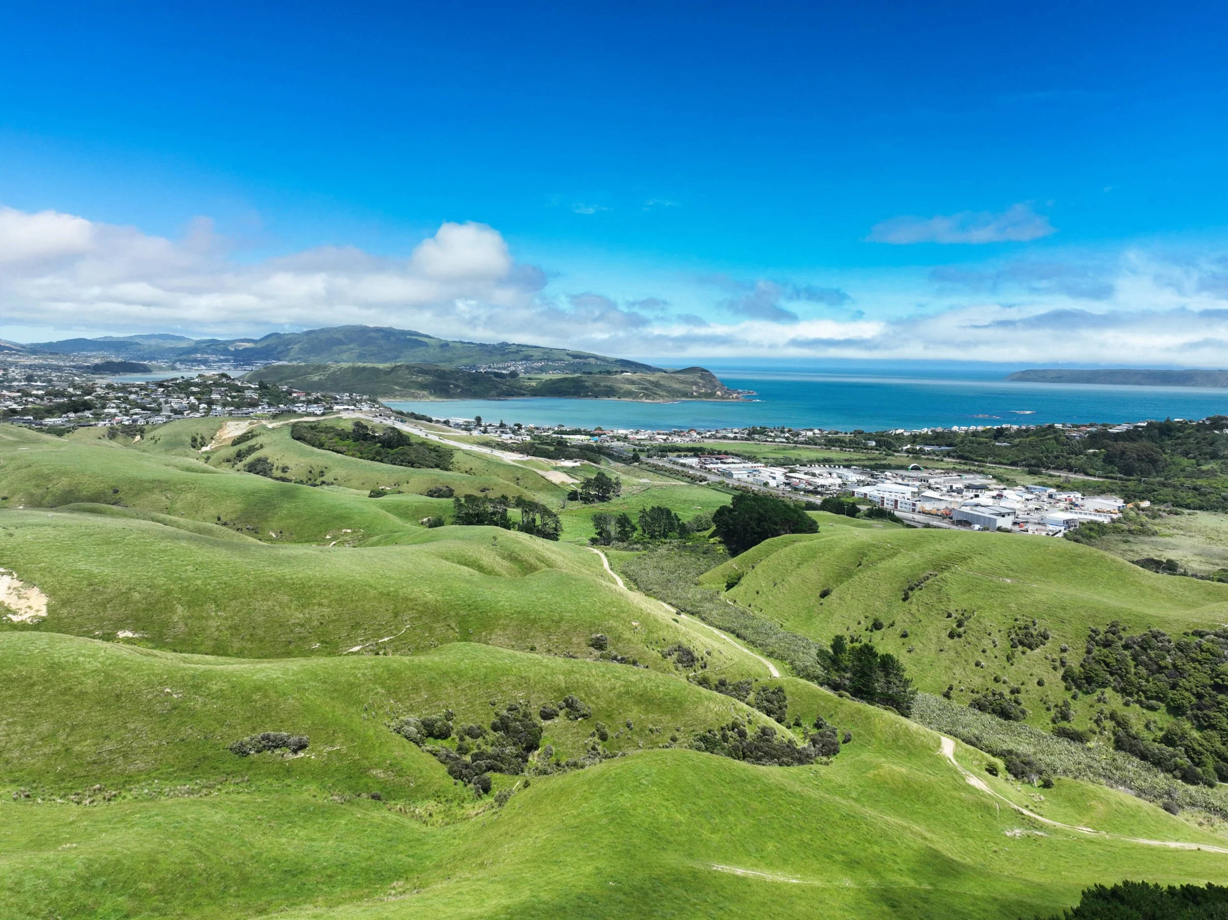 Scenic view of Gillies Group latest development, Plimmerton Farm, and the rolling green hills leading towards a coastal town and bay, with ocean in the background under partly cloudy sky.