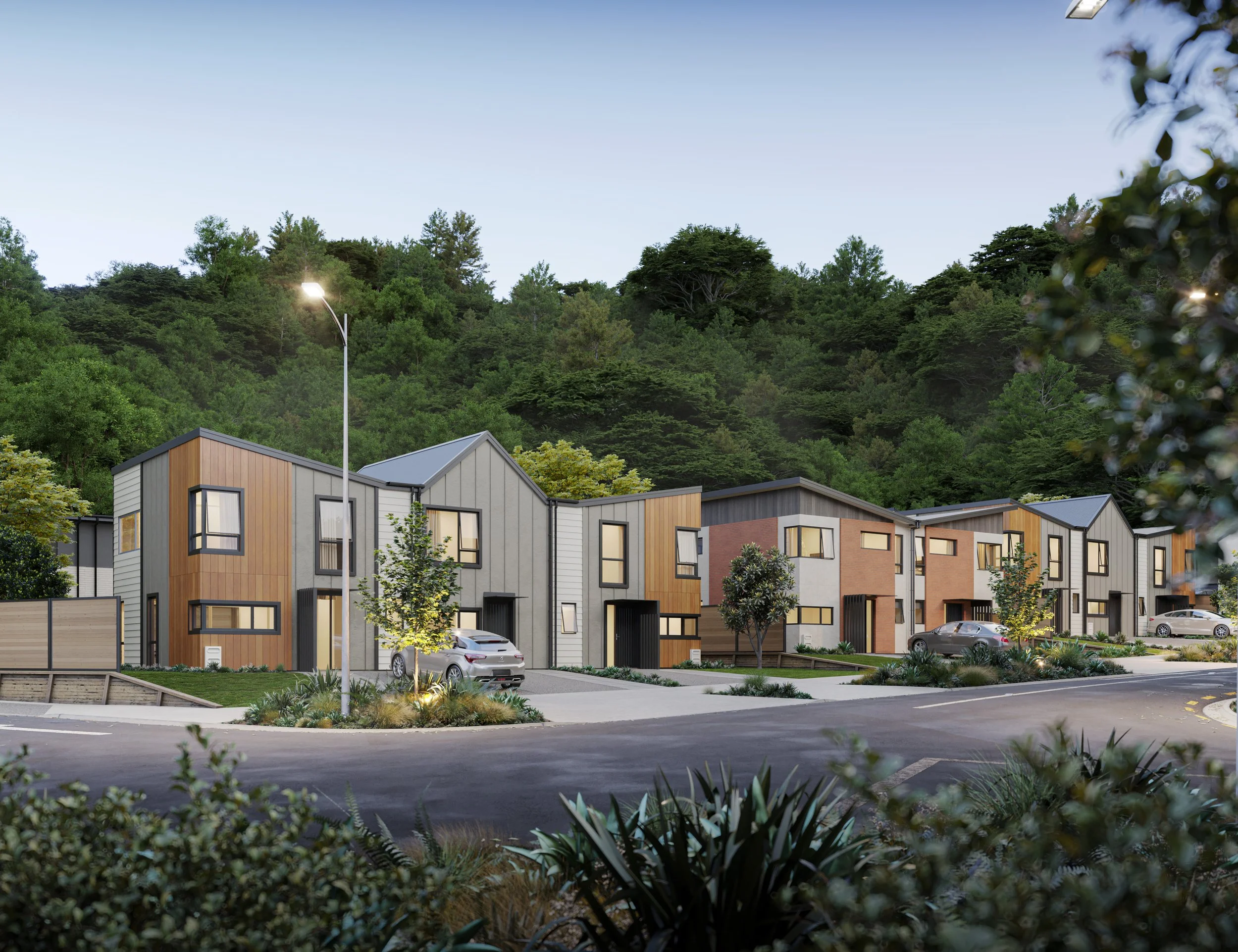 Modern row of townhouses with varied architectural styles, surrounded by landscaped greenery and set against a wooded hillside at dusk.