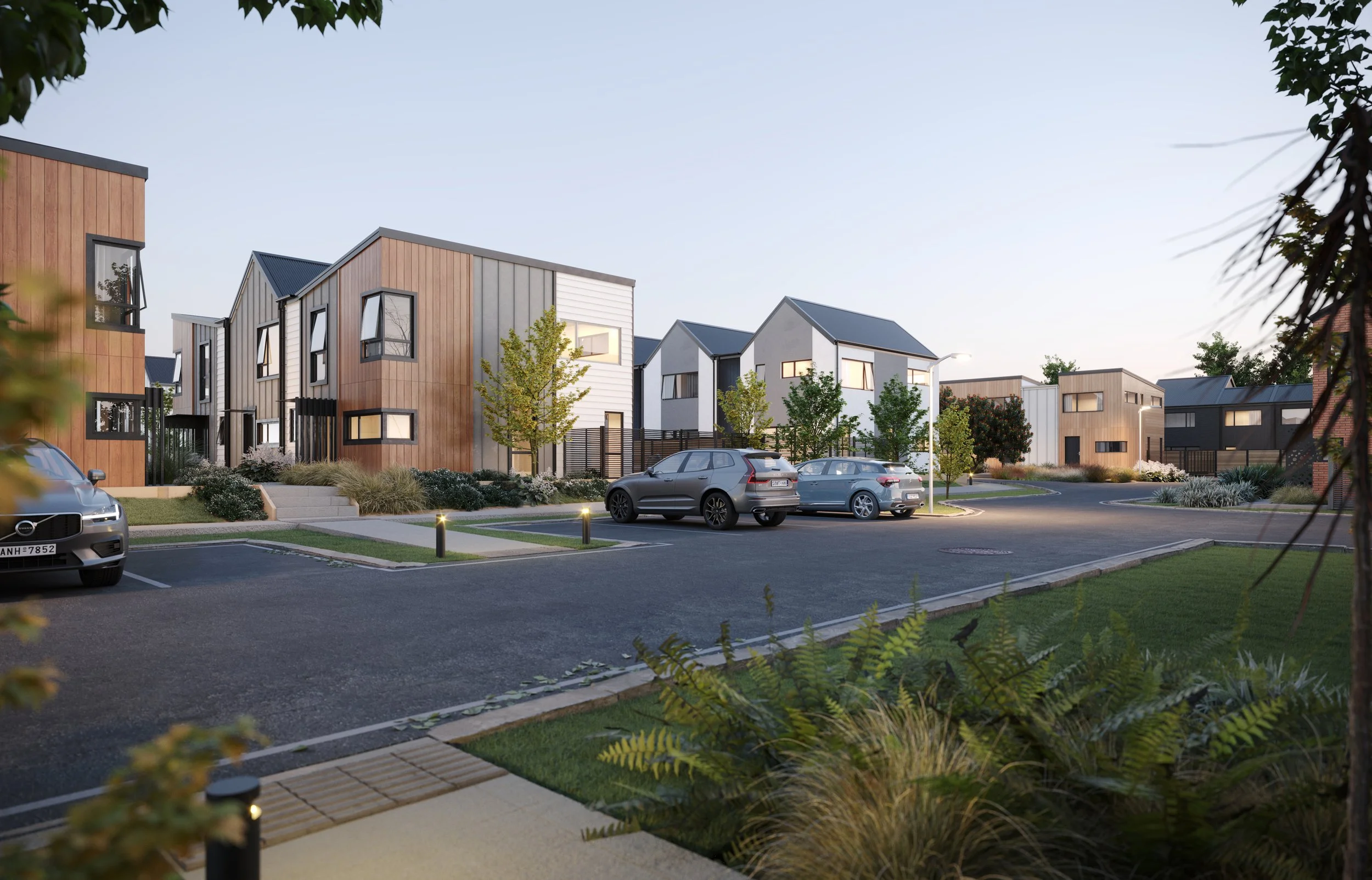 A modern residential community with contemporary two-story houses, parked cars, landscaped greenery, and a paved parking lot during early evening.
