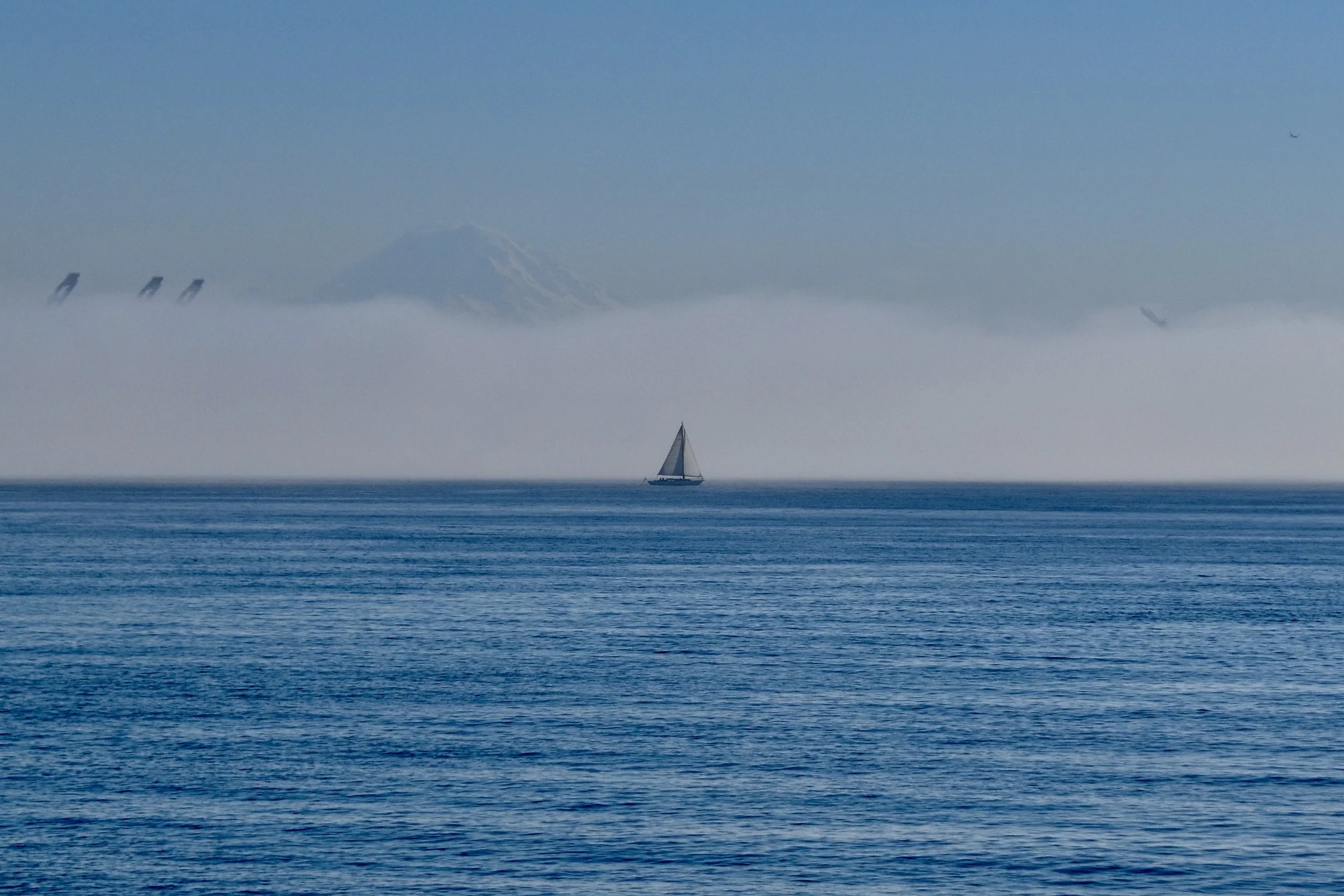 A sailboat on the ocean with Mount Rainier in the background, partially obscured by fog or low clouds, and airplanes flying in the sky.