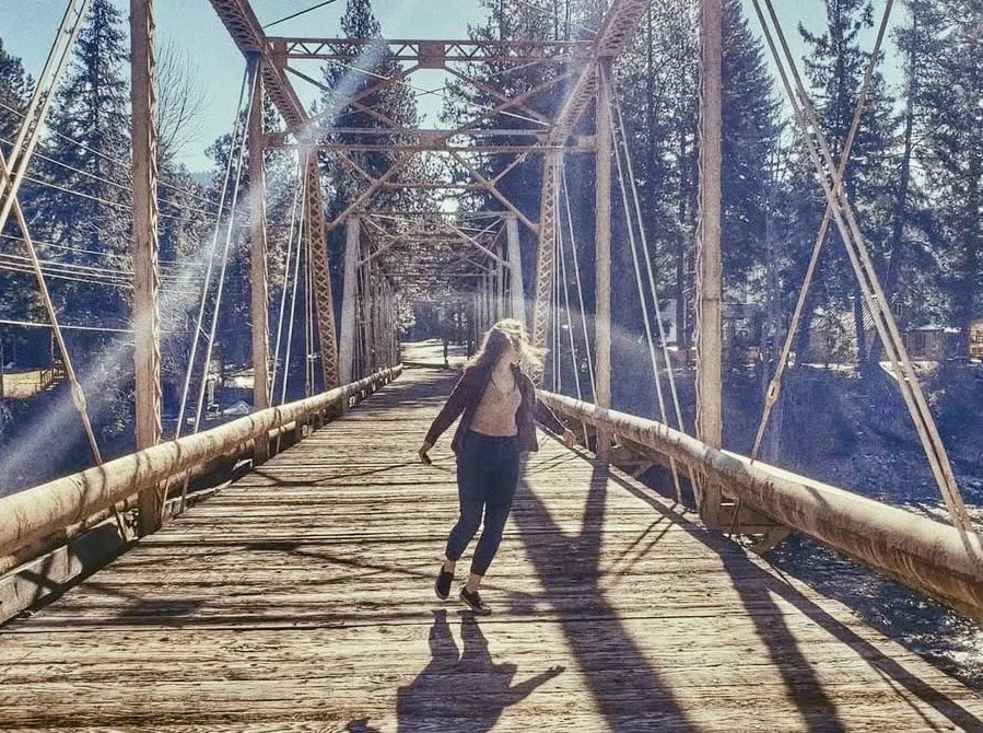 A woman with long brunette hair walking on a wooden suspension bridge surrounded by trees, with sunlight casting long shadows.