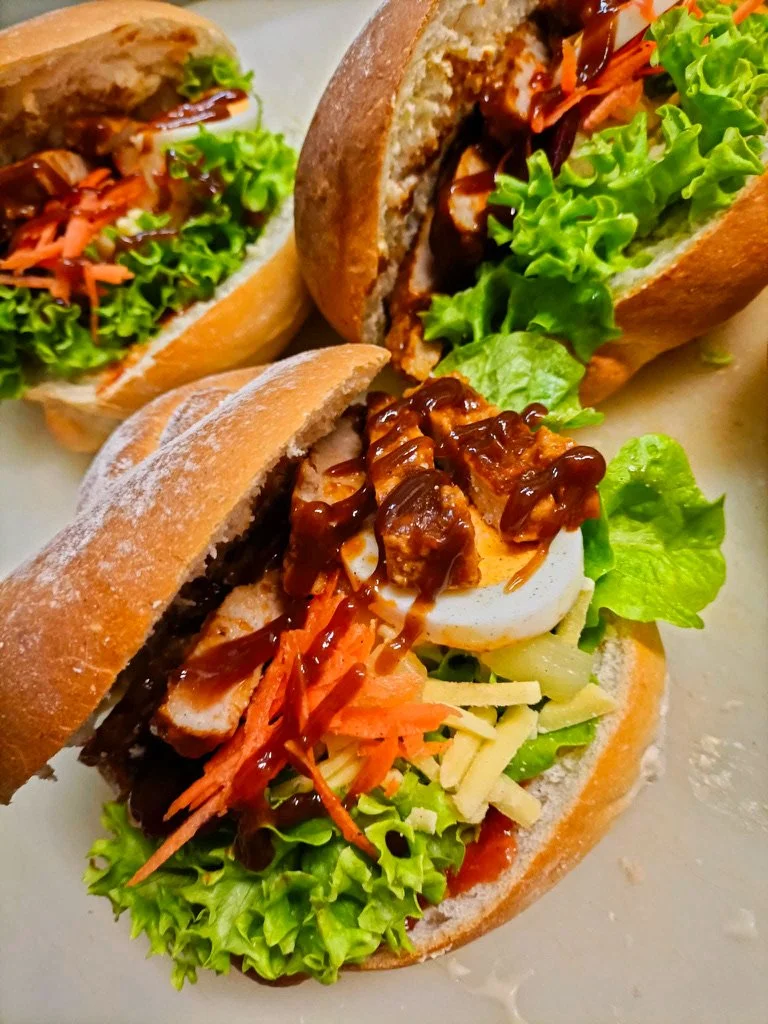Two sandwiches with lettuce, shredded carrots, chicken, shredded cheese, boiled egg, and barbecue sauce in hoagie buns.