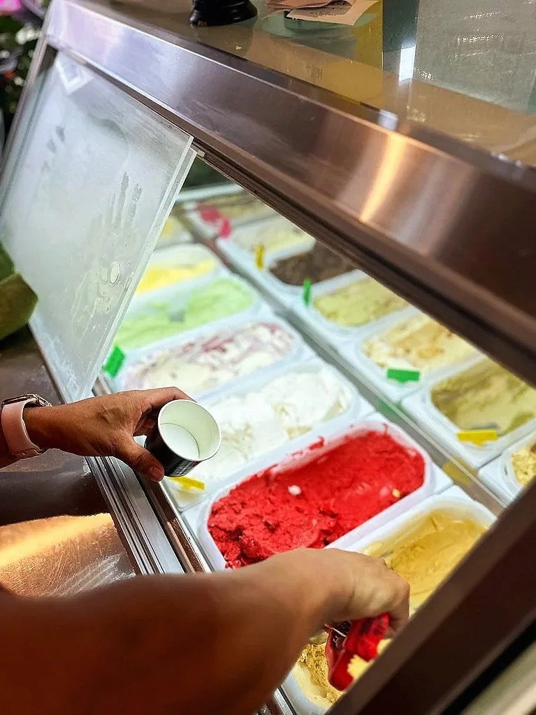 Person scooping red-colored ice cream or gelato from a freezer display case, with various flavors visible.