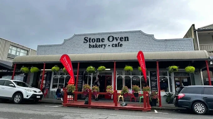 Exterior of Stone Oven bakery and cafe with red flags and potted plants outside, parked cars, and a cloudy sky.