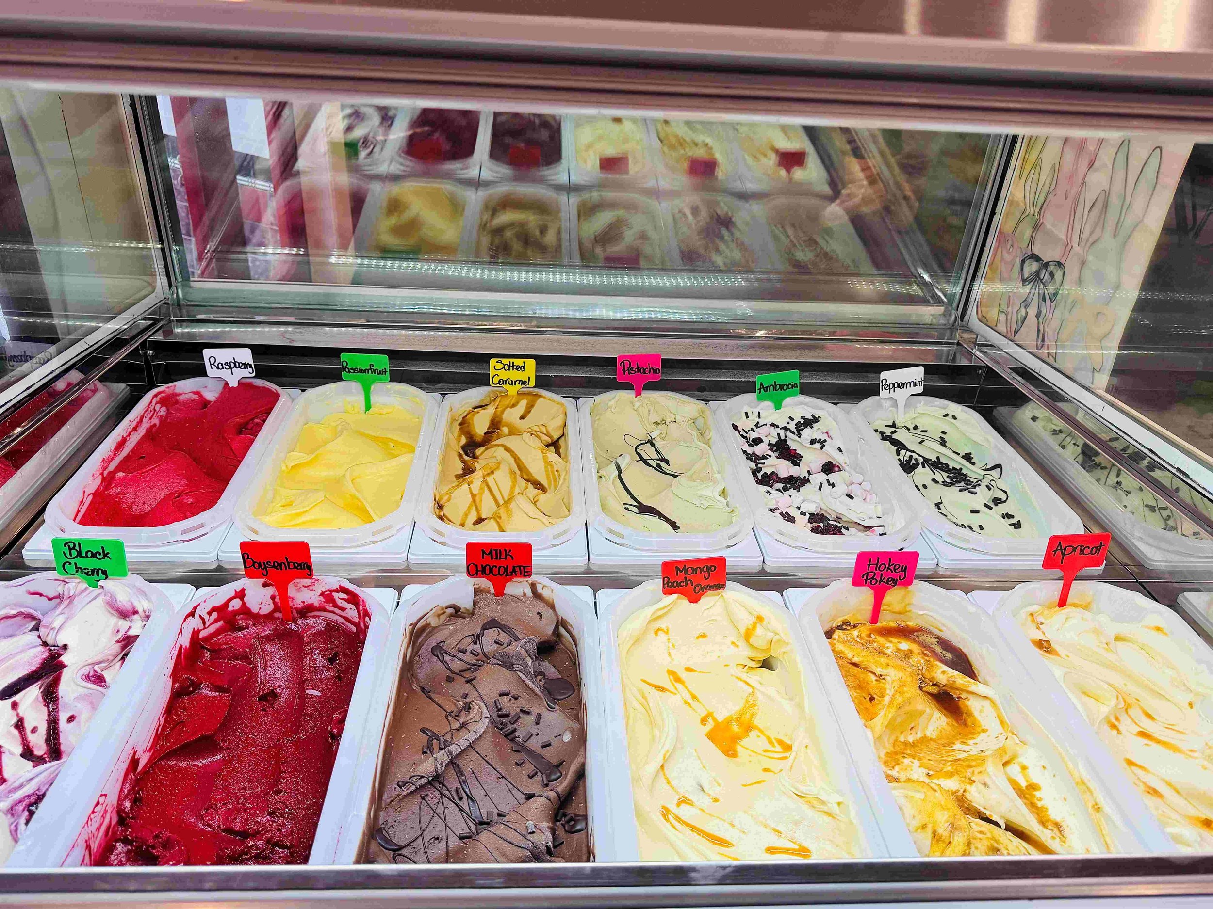 Frozen gelato flavors from stone oven bakery in Hokitika in a display case labeled with colorful signs
