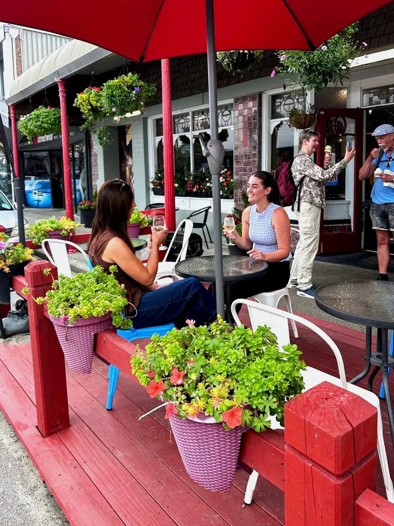 Two women sitting at a table outside a cafe under a large red umbrella, enjoying drinks and smiling at each other outside stone oven bakery & cafe
