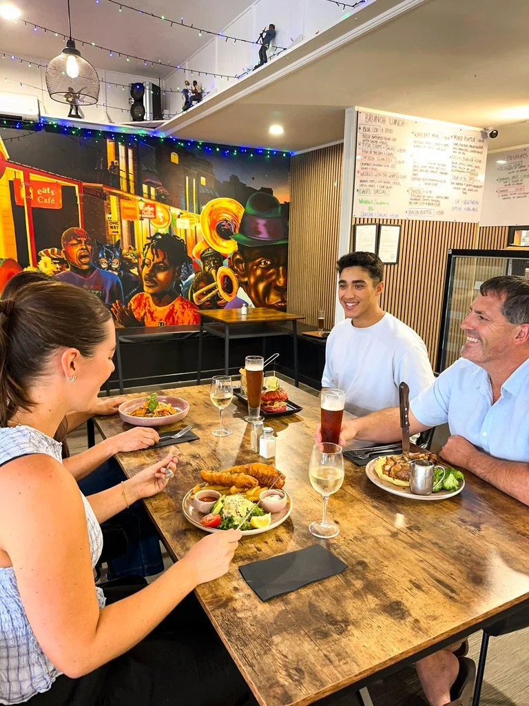 Four people sitting at a wooden table in a restaurant, eating and drinking, with colorful wall art and string lights in the background.