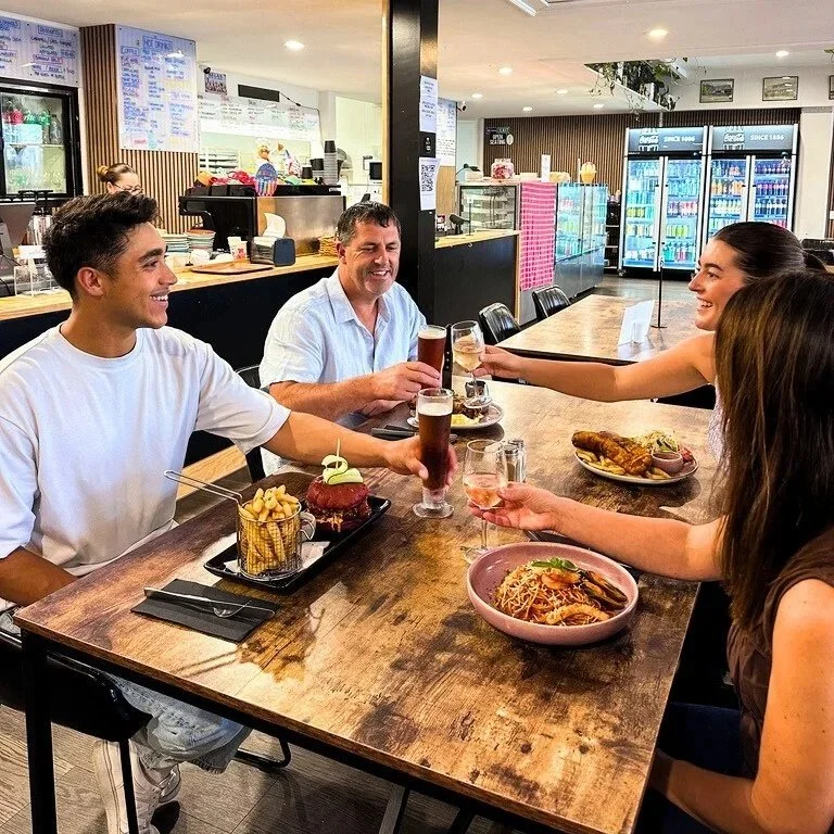 Four friends enjoying drinks and meals at a restaurant bar, clinking glasses in a toast, with various dishes on the table at stone oven bakery for dinner