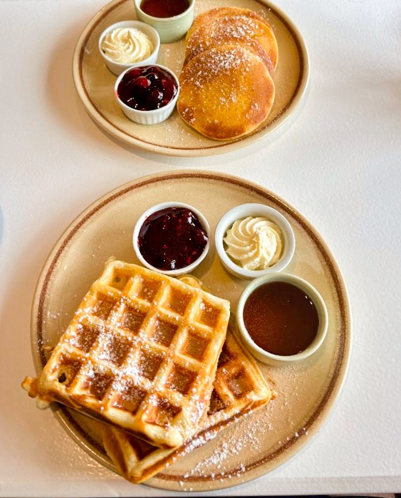 Plate with waffles topped with powdered sugar, served with syrup, whipped cream, and fruit preserves. In the background, another plate has pancakes with powdered sugar, syrup, from stone oven bakery and cafe restaurant in Hokitika