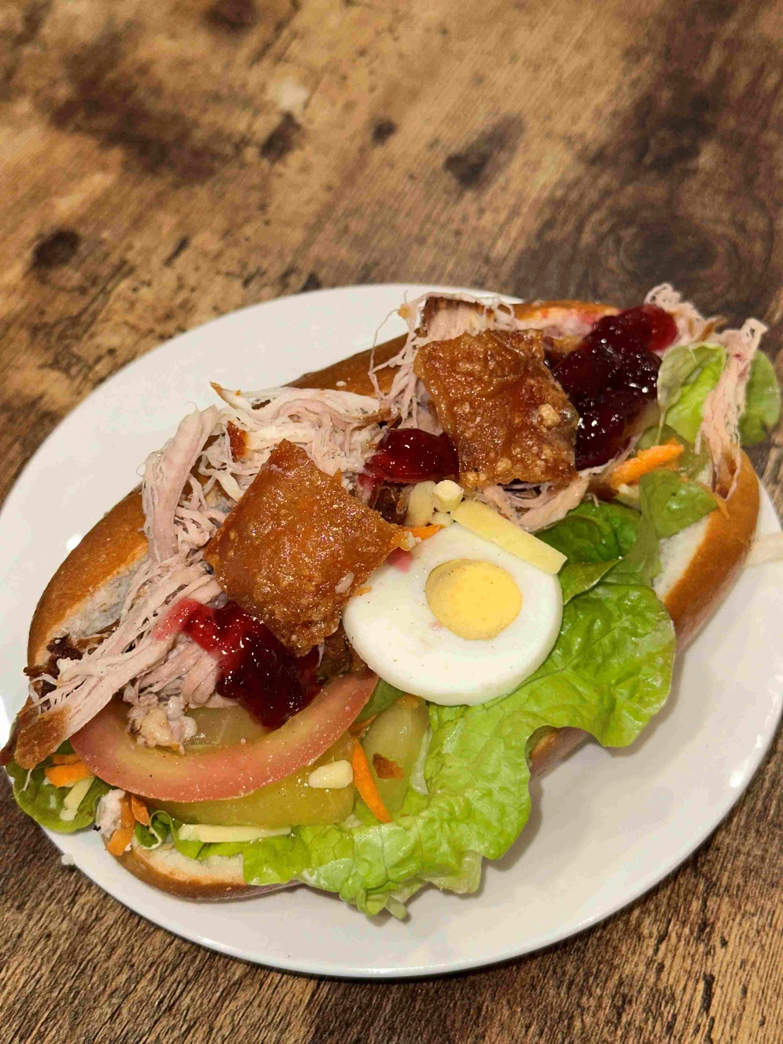 Pulled pork sandwich with lettuce, tomato, pickle, hard-boiled egg, bacon, fried chicken pieces, cranberry sauce, and shredded vegetables on a bun, served on a white plate.