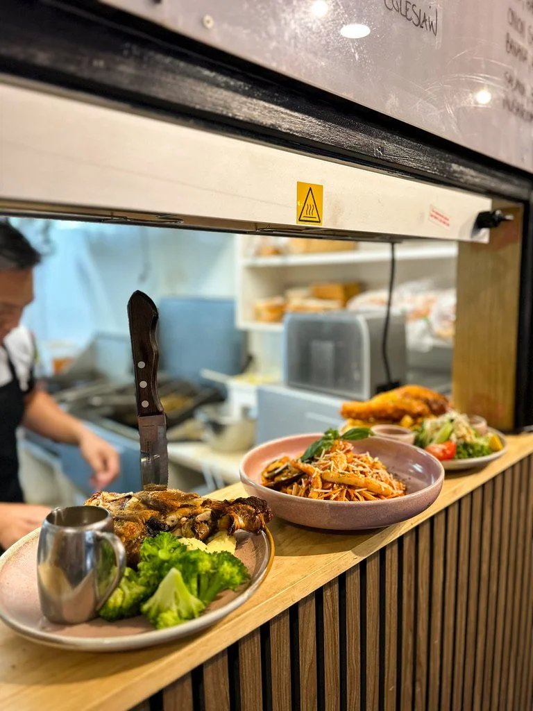 Close-up of a countertop with three plates of food at a restaurant, including grilled chicken, broccoli, pasta with vegetables, and fried chicken with salad, with a kitchen in the background.