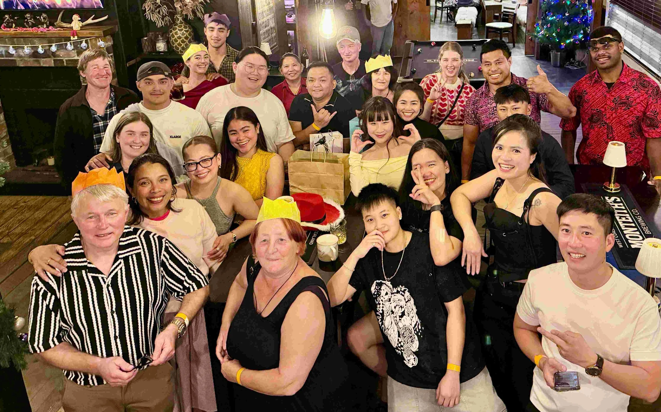 Group of people celebrating at a Christmas party, with some wearing paper crowns, a Christmas tree, and festive decorations in the background.