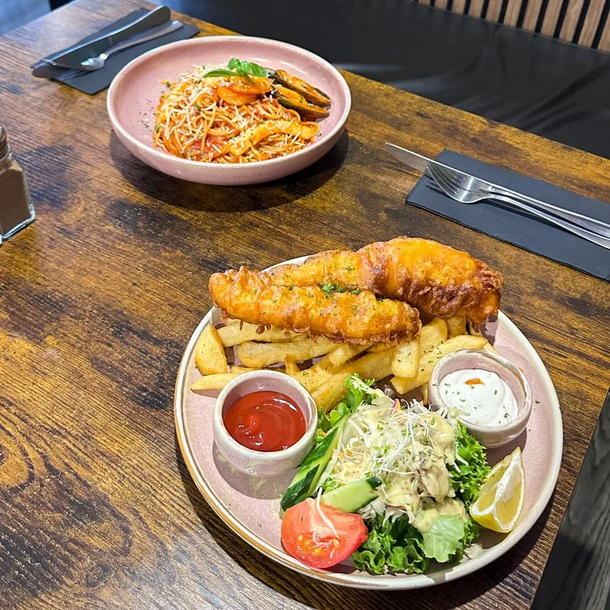 Plate of fish and chips with sides of fresh salad and condiments, and a bowl of pasta on a wooden table in a restaurant setting.