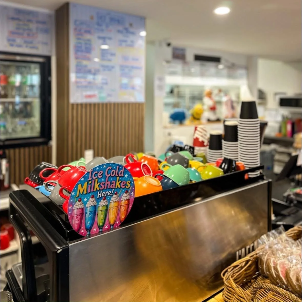 Colorful coffee mugs and paper cups on a counter in a coffee shop, with a sign advertising ice cold milkshakes.