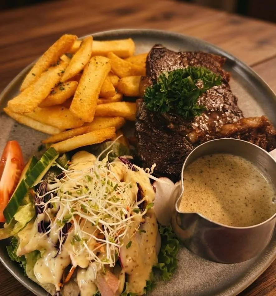 A plate of food with French fries, a grilled steak garnished with parsley, a side salad with sprouts, cucumber, and tomato, and a cup of peppercorn sauce.