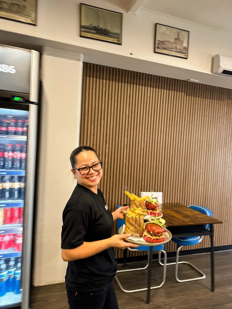 A woman with glasses and dark hair pulled back, smiling while holding a tray with sandwiches and fries, standing in a restaurant or cafe with a wooden wall, tables, and a vending machine in the background.