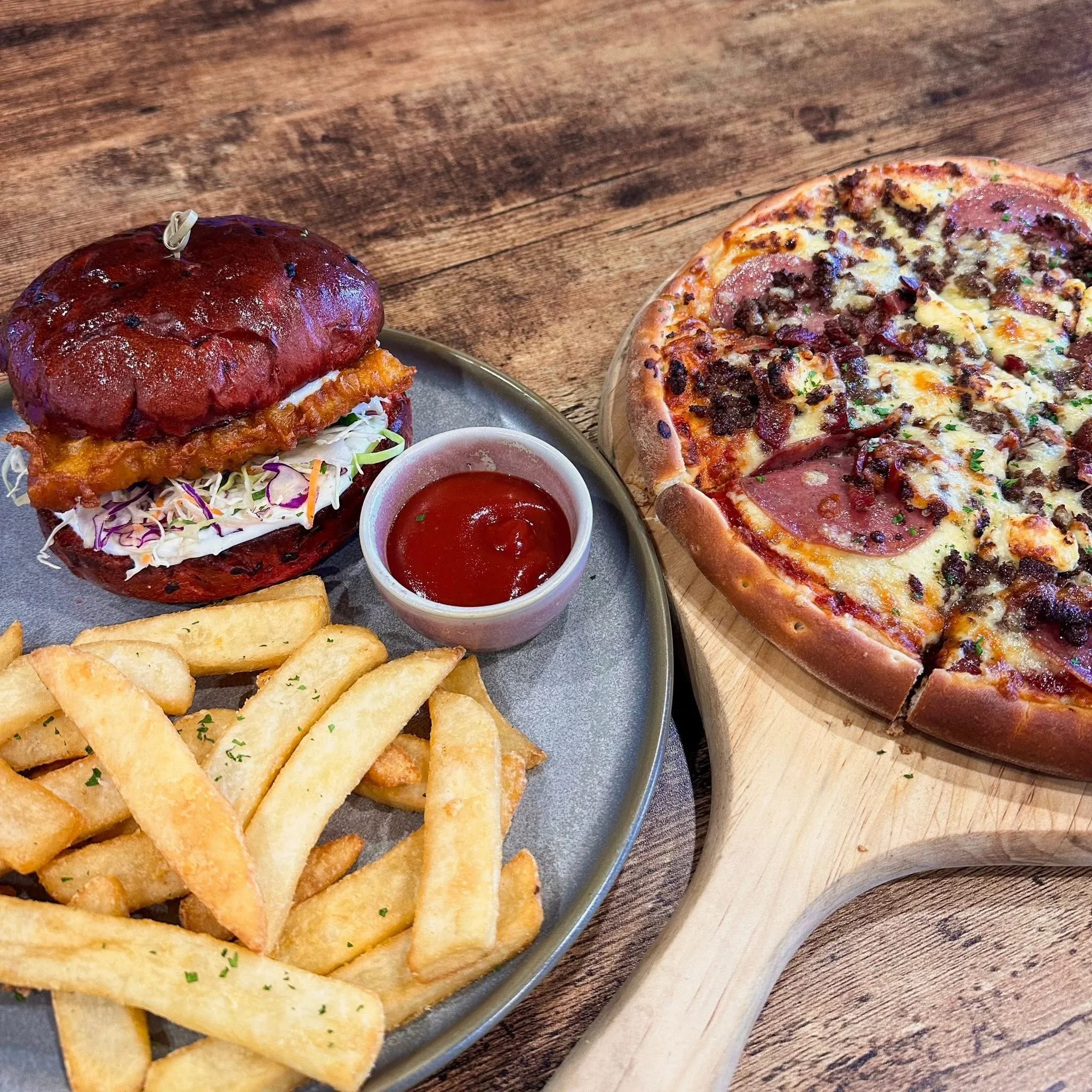 A plate with a fried chicken sandwich with coleslaw, fries, and a cup of ketchup, alongside a pepperoni, sausage, and bacon pizza on a wooden table.