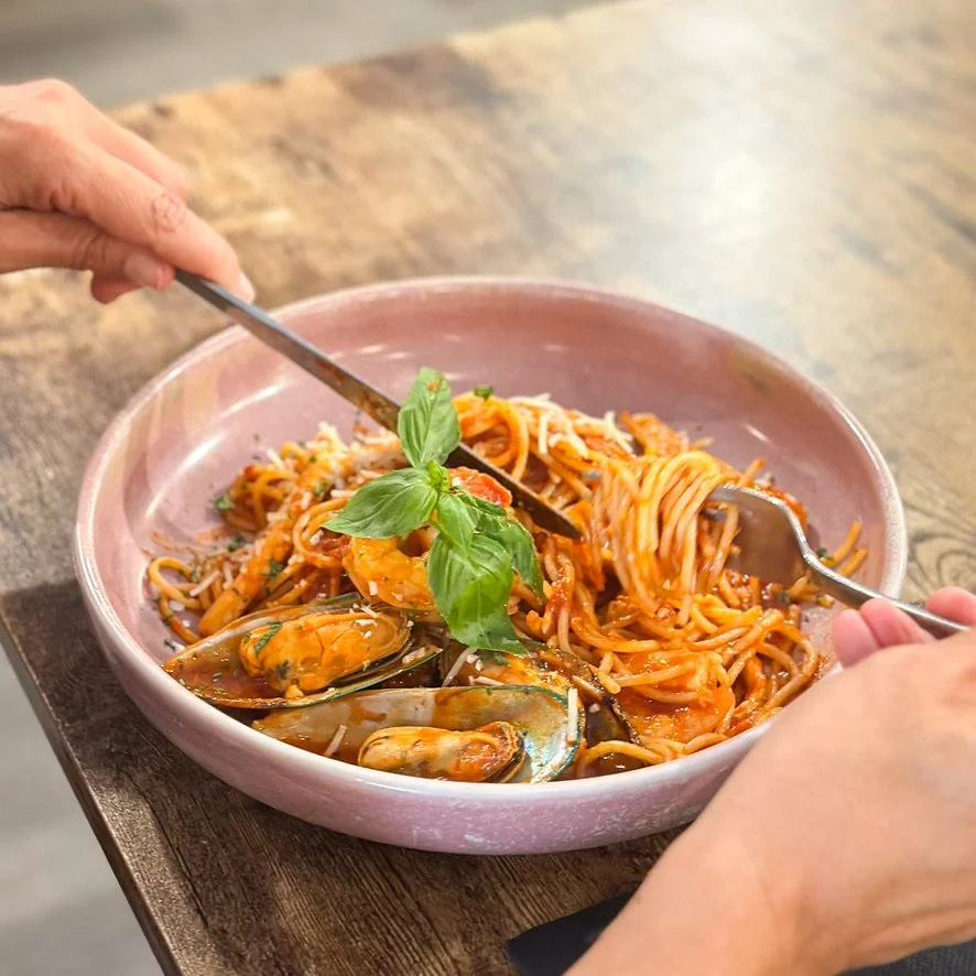 A pink bowl of seafood spaghetti with mussels, shrimp, and basil from stone oven bakery and cafe restaurant dinner menu in Hokitika