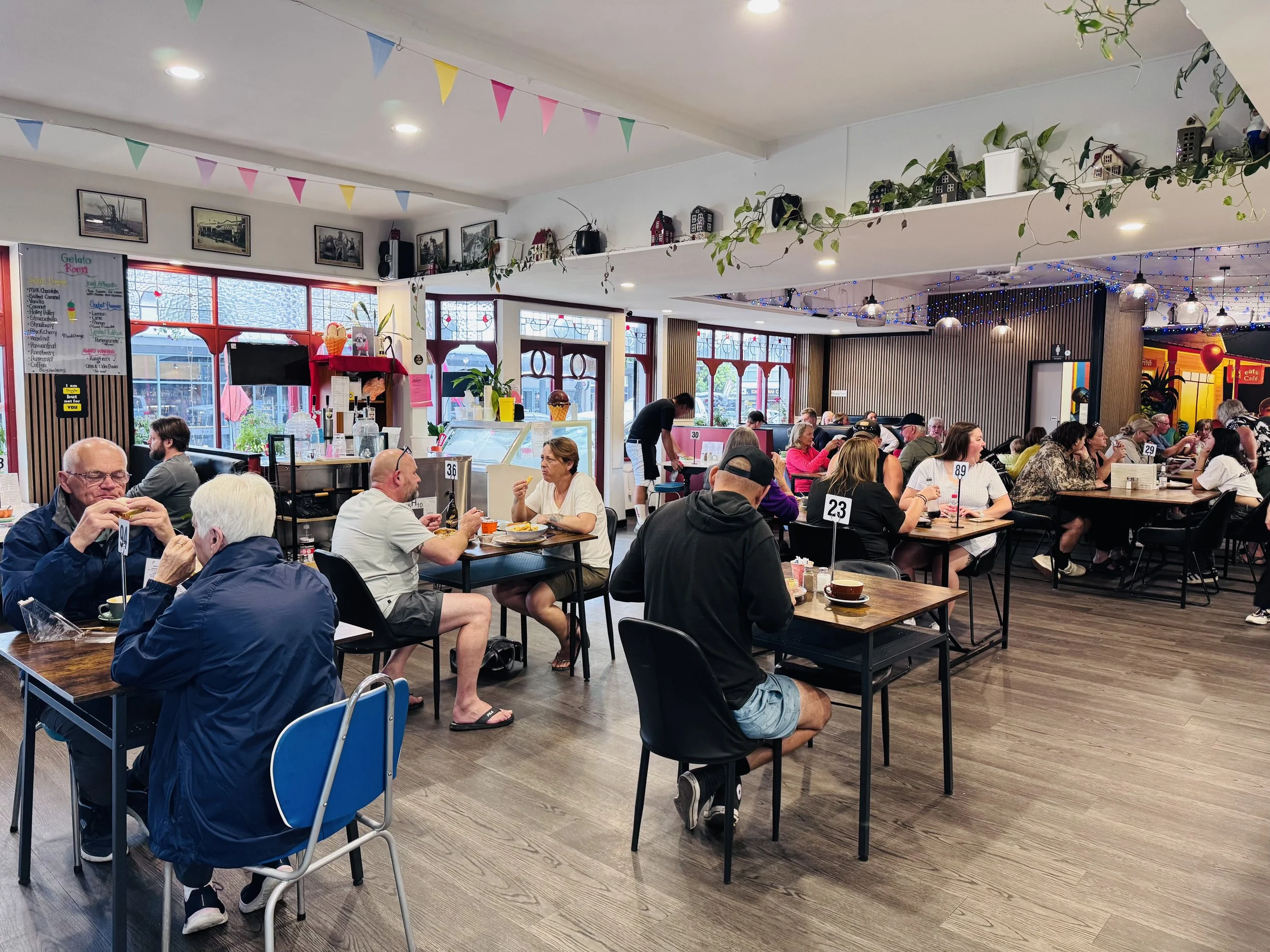 Inside a busy restaurant with multiple tables filled with customers eating and talking. Bright lighting, colorful decorations, and large windows with red frames. Wall art, plants, and string lights enhance the lively atmosphere.