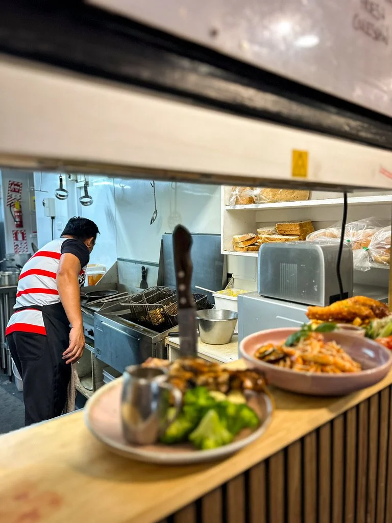 A restaurant kitchen with a chef cooking behind a counter, with plates of food and fresh ingredients in the foreground.
