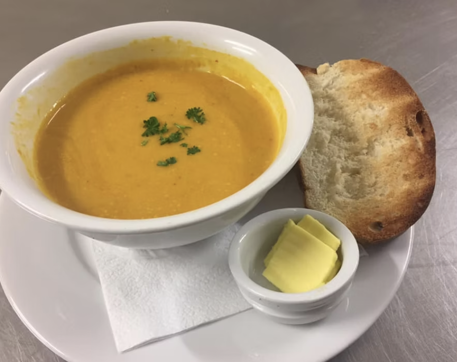 A bowl of orange lentil soup garnished with cilantro, served with a piece of toasted bread and a small dish of butter on a white plate.